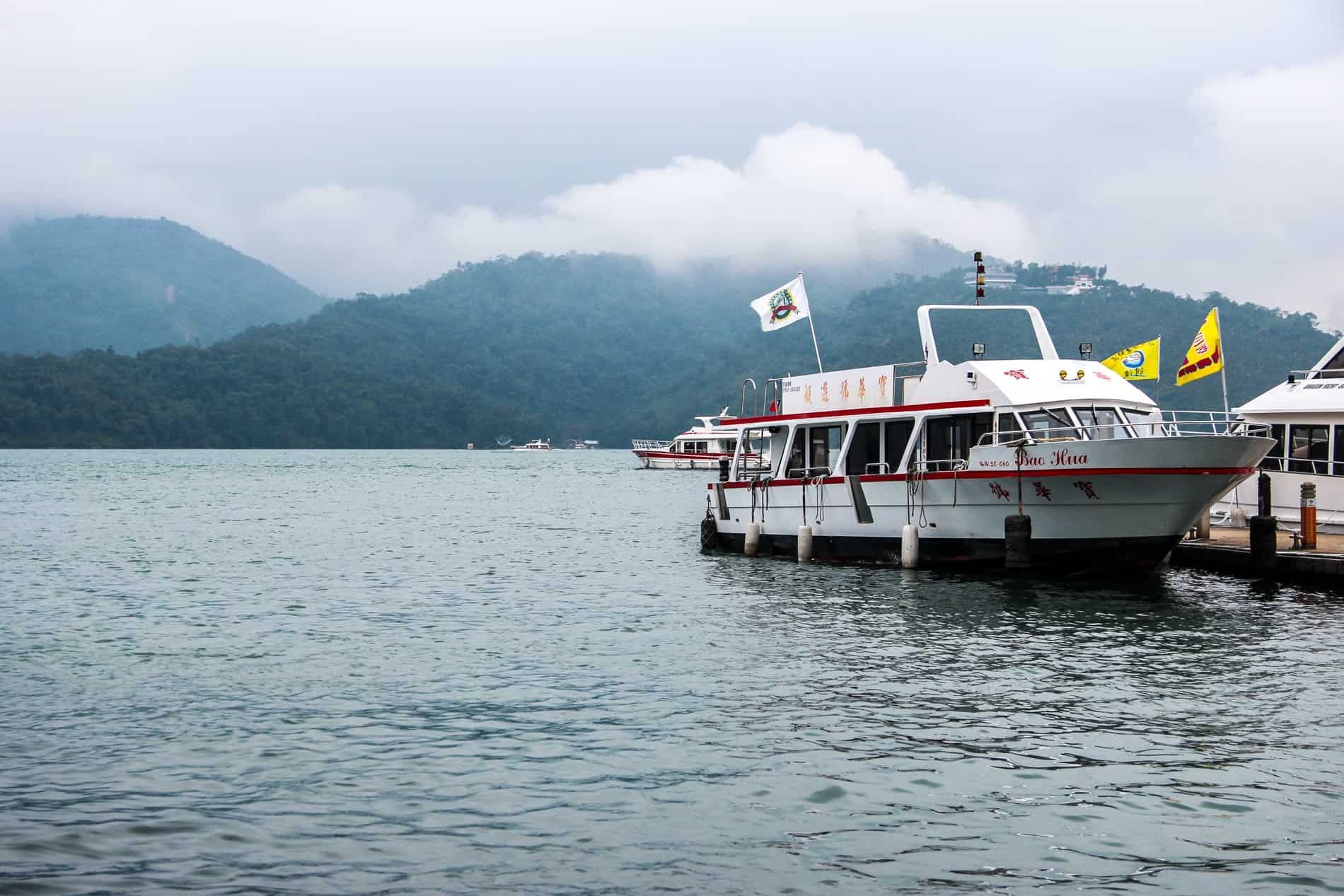 A white boat docks at a small pier on the large expanse of mountain backed water in Taiwan. 