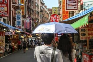 A couple under a blue umbrella walking through a busy street market in Taipei.