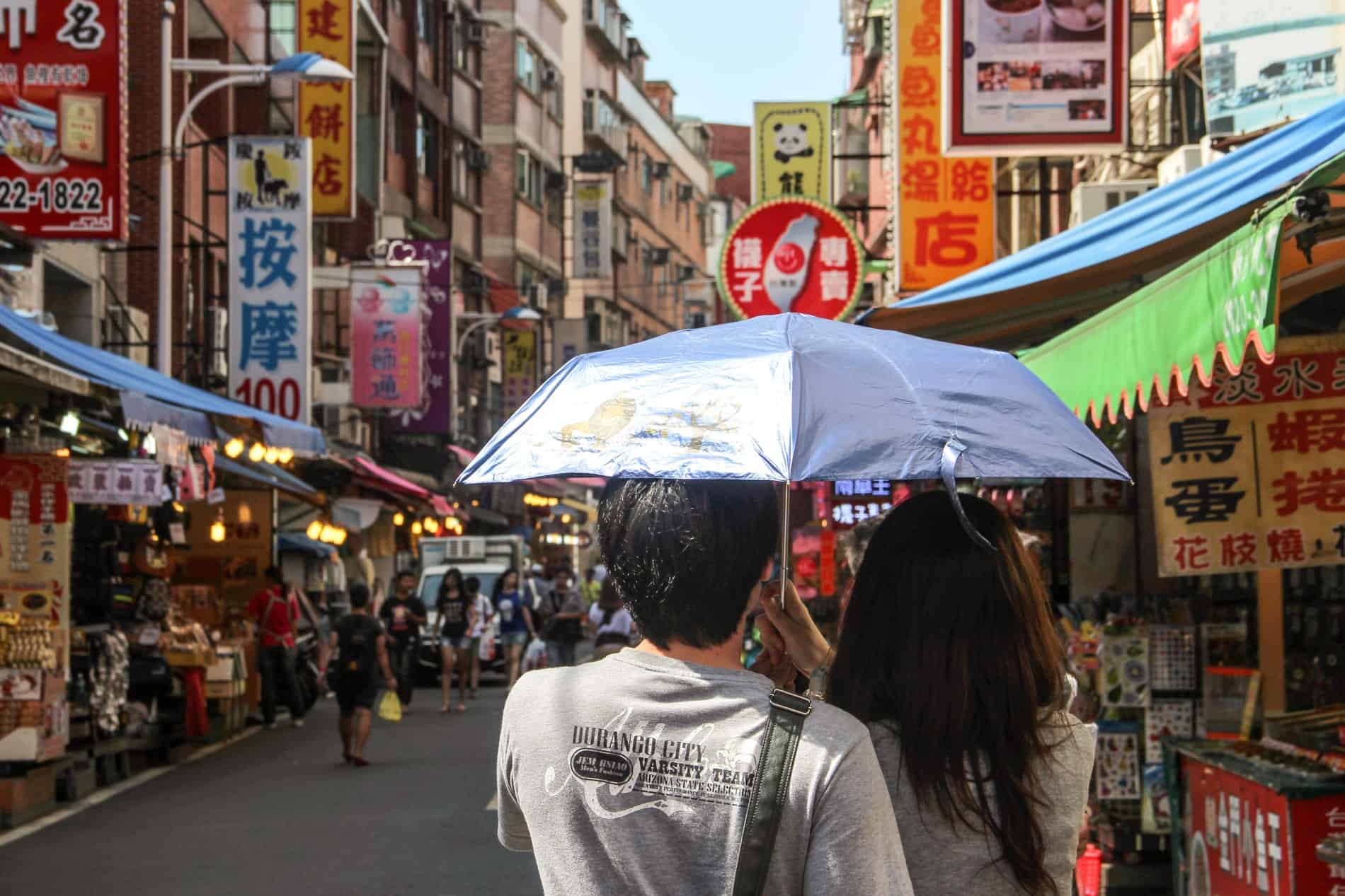 A couple under a blue umbrella walking through a busy street market in Taipei. 