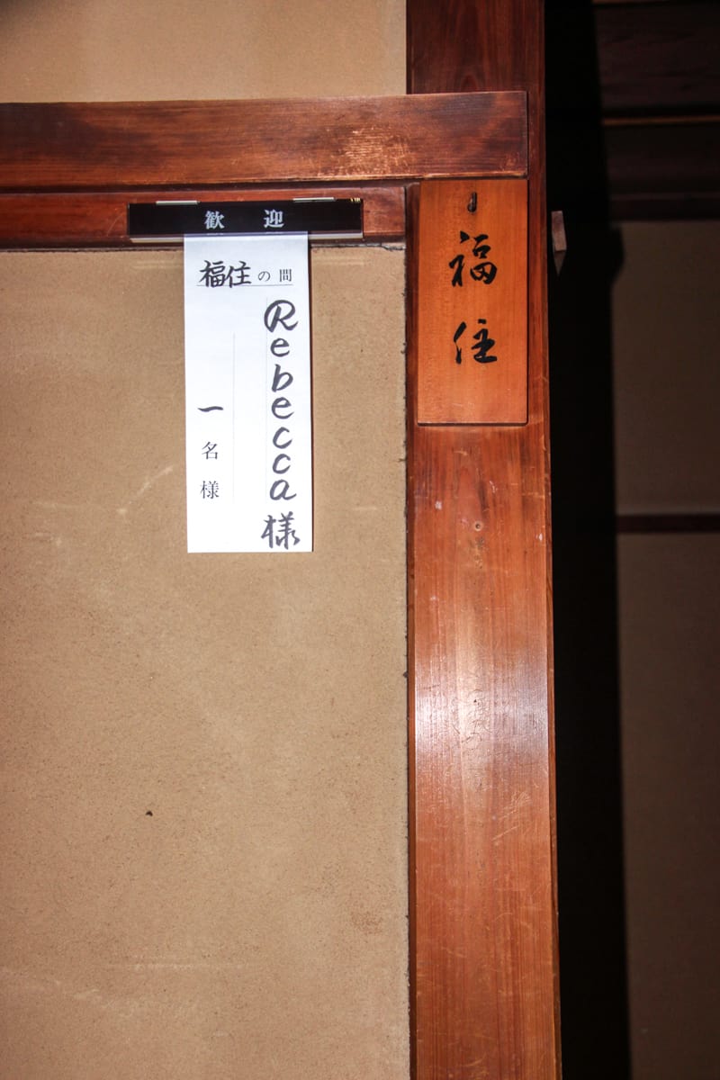 A wooden door with a white paper sign in the corner which has Japanese characters and the name 'Rebecca' written in black pen.