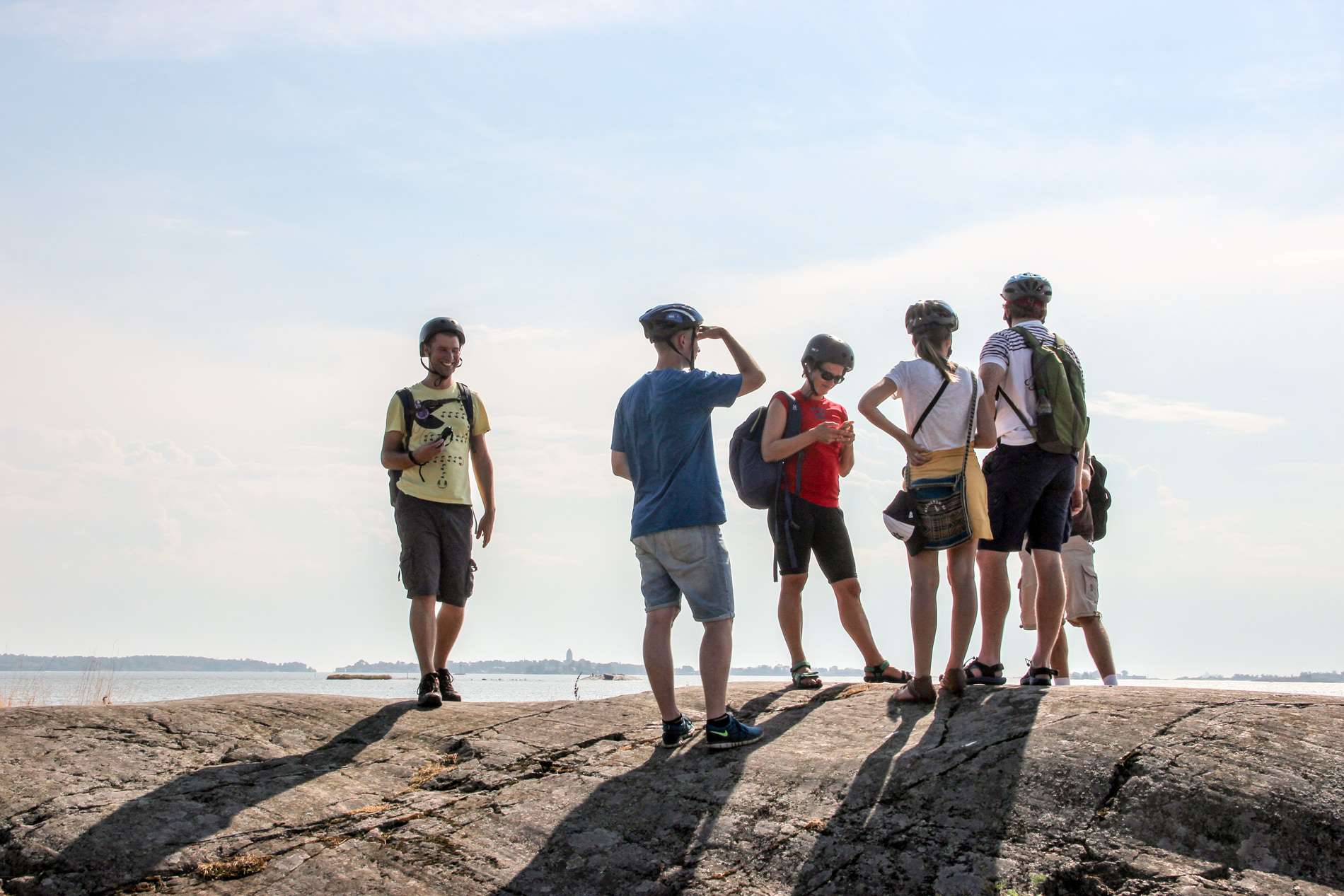 Six people in helmets sand upon a large rock looking out to sea in Helsinki. 