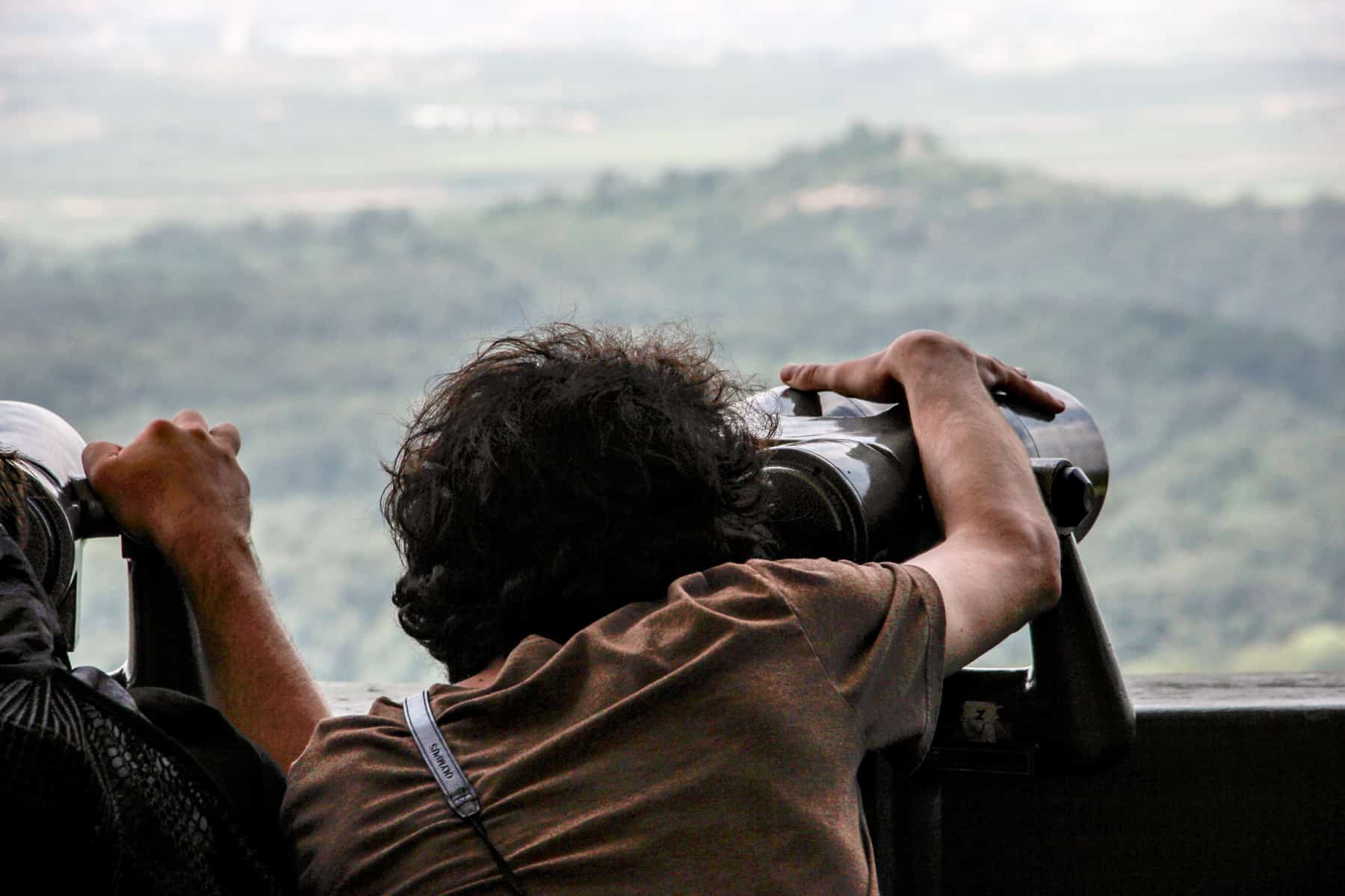 A woman in a brown t-shirt looks through a huge set of binoculars towards a green mound - the Propaganda Village in North Korea, seen on a trip to the DMZ.