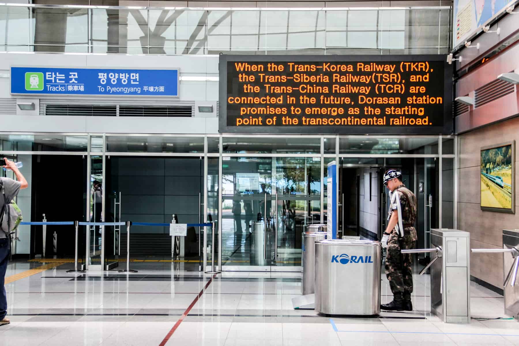 A South Korean military guard at the entrance barrier at Dorasan Train Station, seen on a DMZ tour. A blue sign signals the direction to Pyeongyang (the capital of North Korea) and a black sign has orange text reading "When the Trans-Korea Railway (TKR), the Trans-Siberia Railway (TSR), and the Trans-China Railway (TCR) are connected in the future, Dorasan station promise to emerge as the starting point of the transcontinental railroad. 