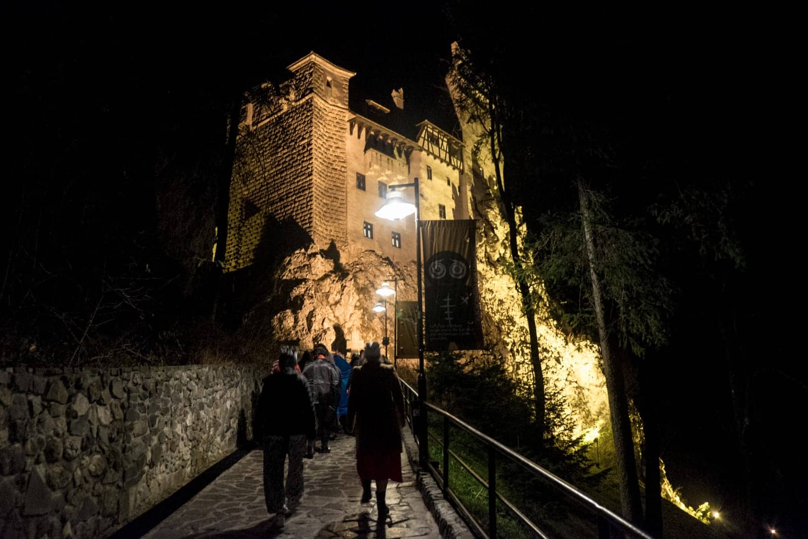 A group of people walk towards Bran Castle in Transylvania, Romania at night. It's lit with a golden light. 