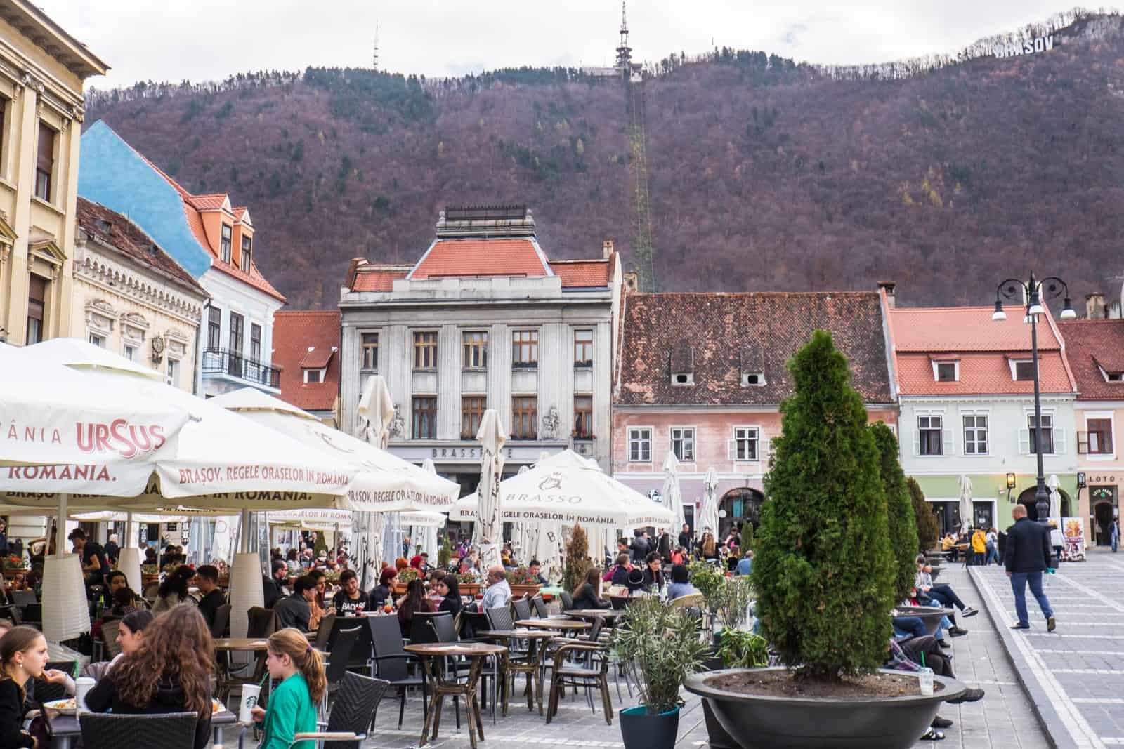 Groups of people enjoying al freso dining in Brasov city main square in Romania. The pastel hued building with red rooftops are backed by high hills. 