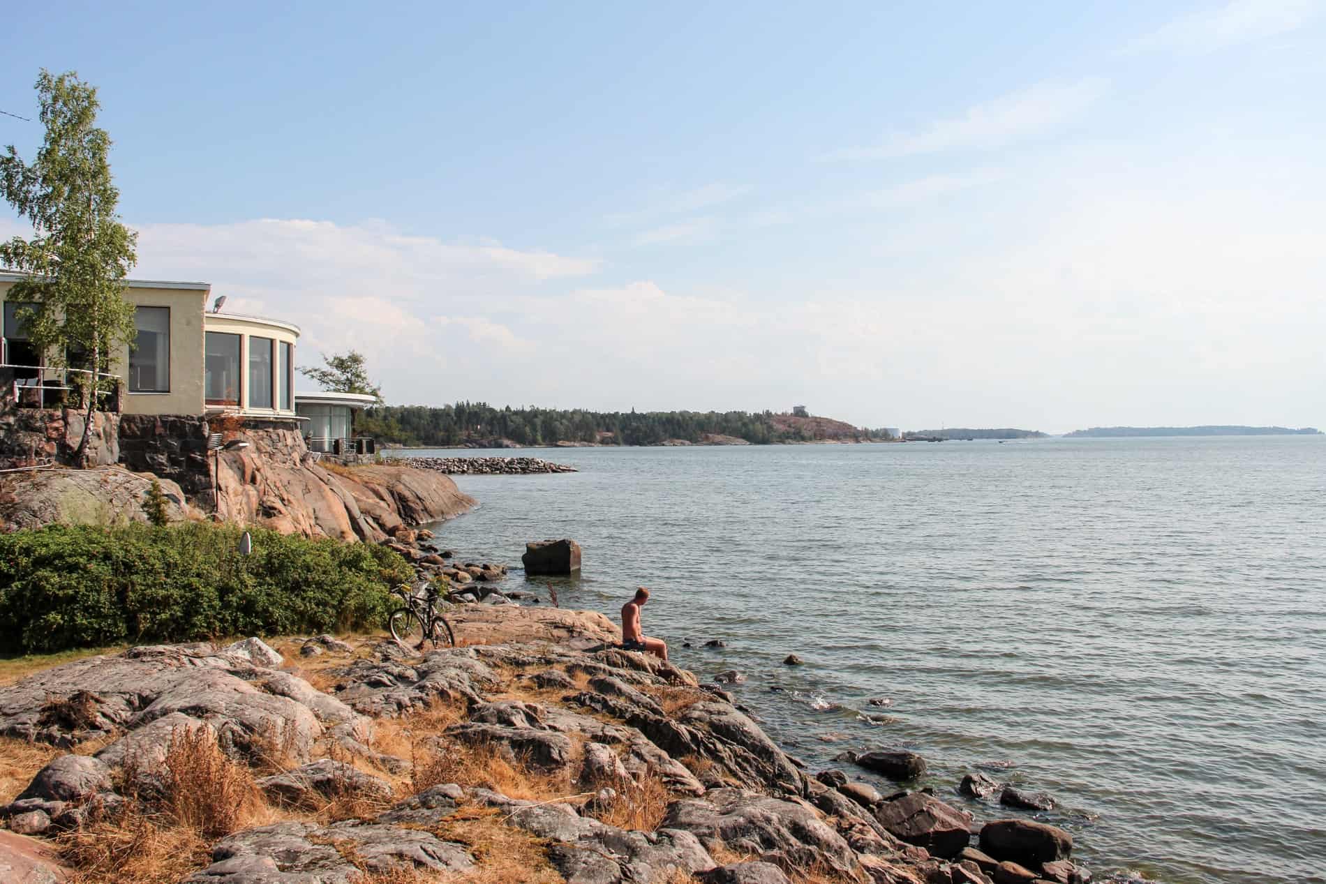 In front of a house, a man sits on a rocky mound on the coastline of Marjaniemi beach in Helsinki. 
