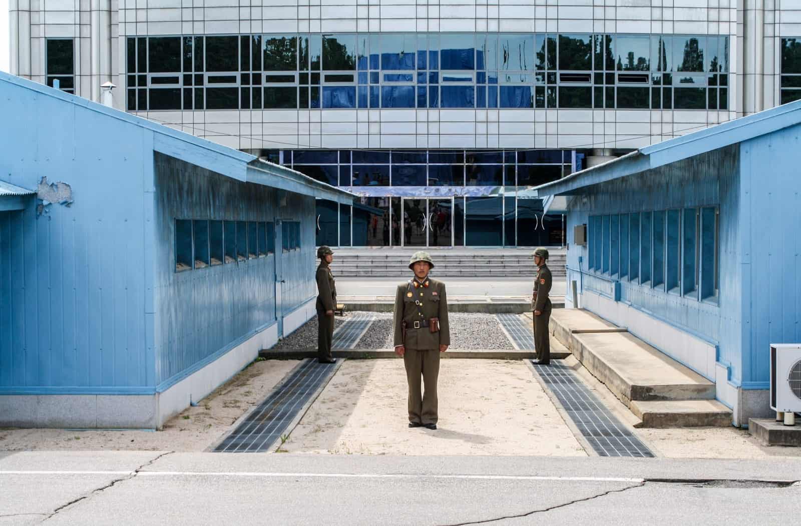 Three North Korean guards in green uniform stand between two blue huts on the North South Korean border at the DMZ demiliterized zone on a DMZ tour.