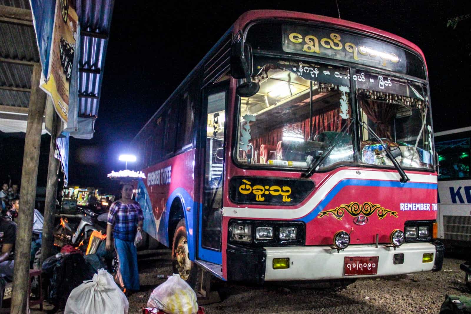 A red Myanmar night bus waits at the dusty station. Tied bags of rubbish stand to the left of the bus, as a woman walks buy carrying a plate of goods on her head. 