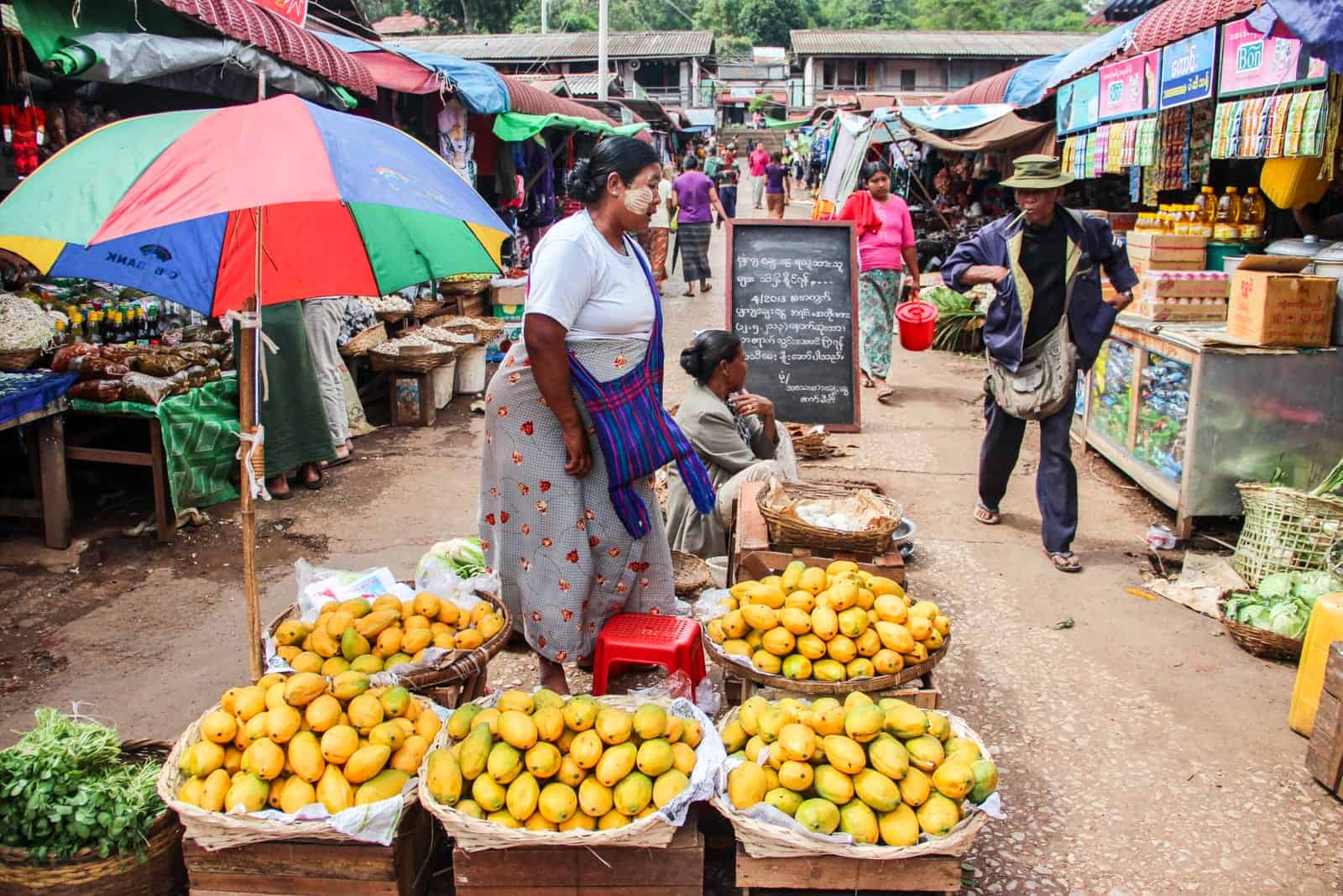 A woman wearing a purple bag by her stall in a local market in Myanmar, with five stands of bright yellow mangoes and a red-blue-green-yellow umbrella. Rows of stalls on either side of the street can be seen in the background.