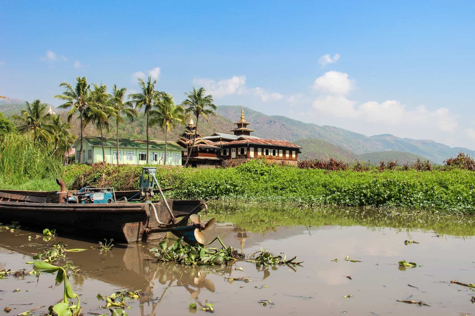A rusting boat with a blue engine, sits in a pool of murky water, backed by green water plants. In the far distance is a red temple with a golden pagoda in the countryside of Myanmar, next to a mint green building and a row of six palm trees. 