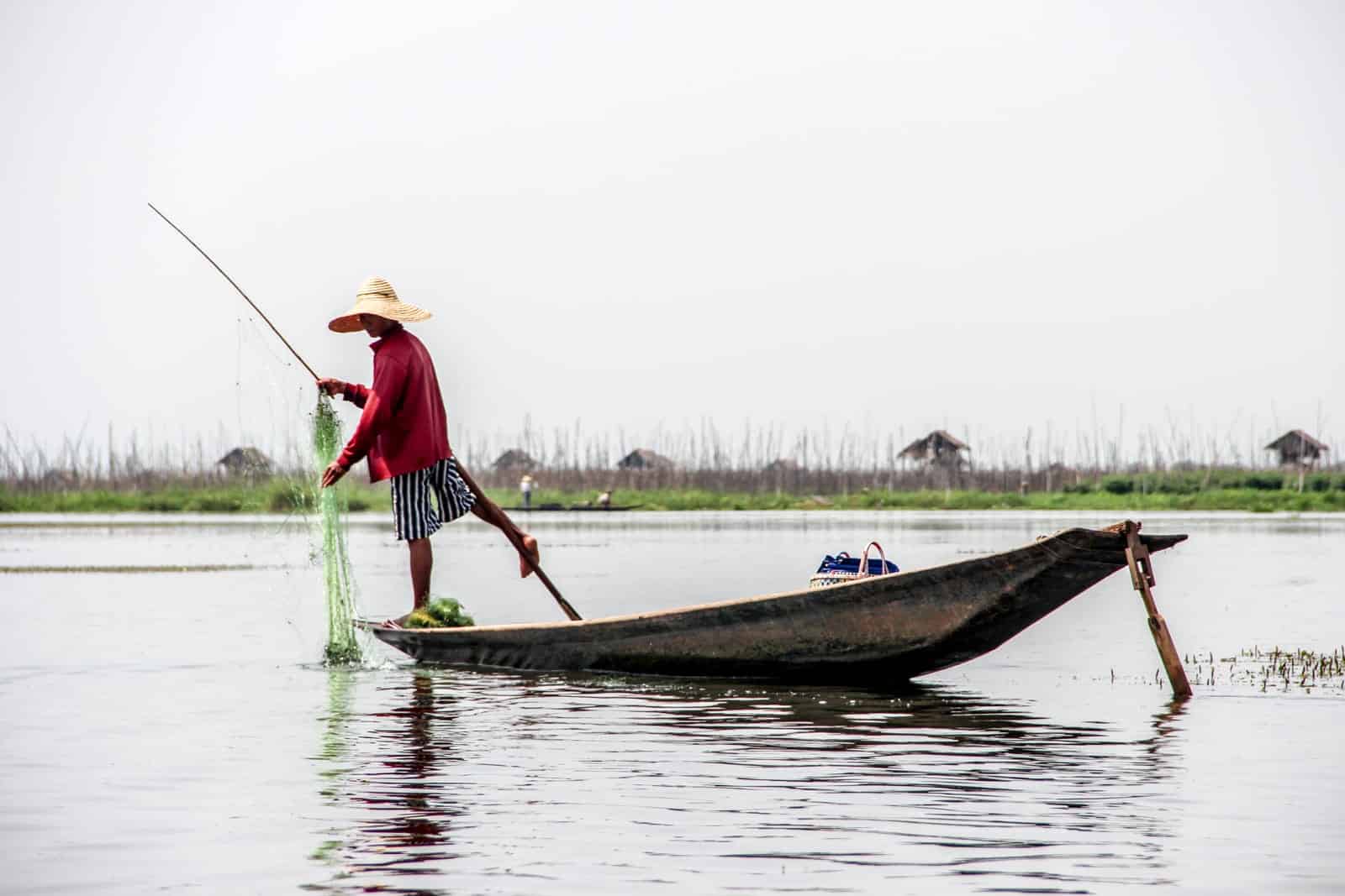 A fisherman in a red top, stripy shorts and a straw hat on Inle Lake in Myanmar practicing the traditional method of rowing the oar with one leg