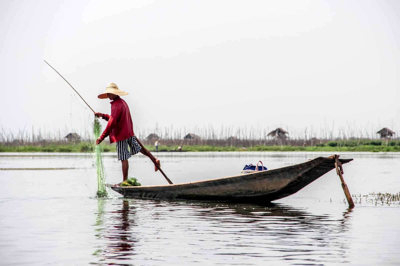 A fisherman in a red top, stripy shorts and a straw hat on Inle Lake in Myanmar practicing the traditional method of rowing the oar with one leg.