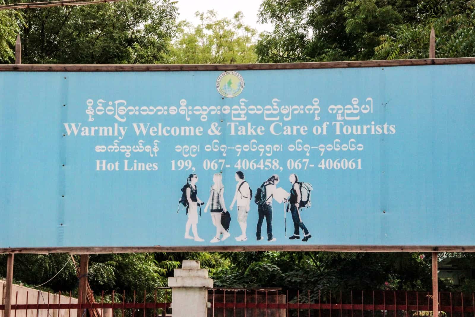 A sky blue coloured sign in Myanmar stating "Warmly Welcome & Take Care of Tourists" with a hotline number 