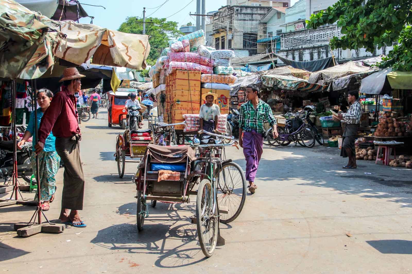 A man in a red shirt, beige hat and green wrap skirt stands next to a market stall while a man in a green shirt and purple tunic and a bike packed high with boxes pass. A bustling street scene in Myanmar.