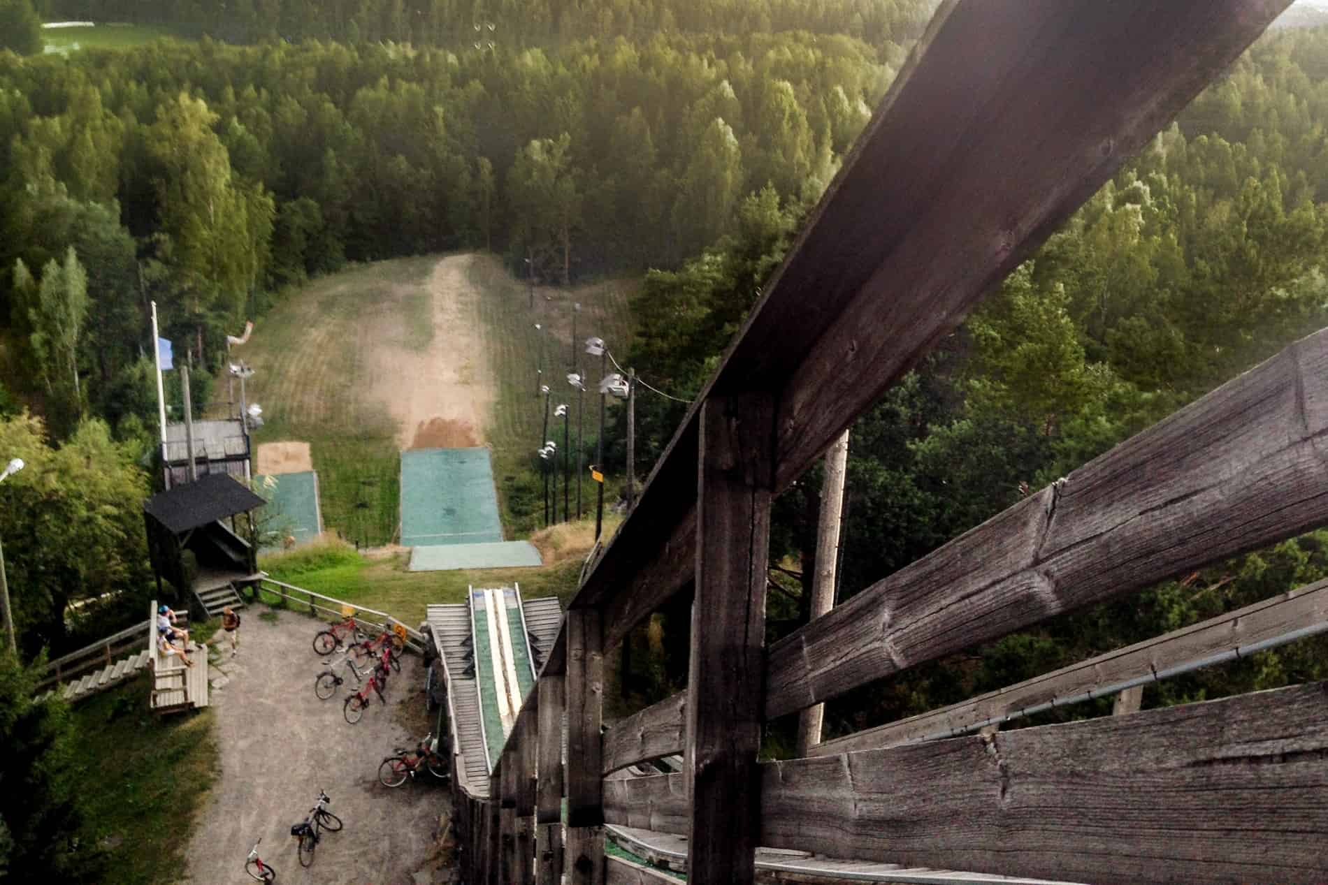 View from the top of a ski jump in summer season looking down towards the forest-set landing point and a group of biks. 