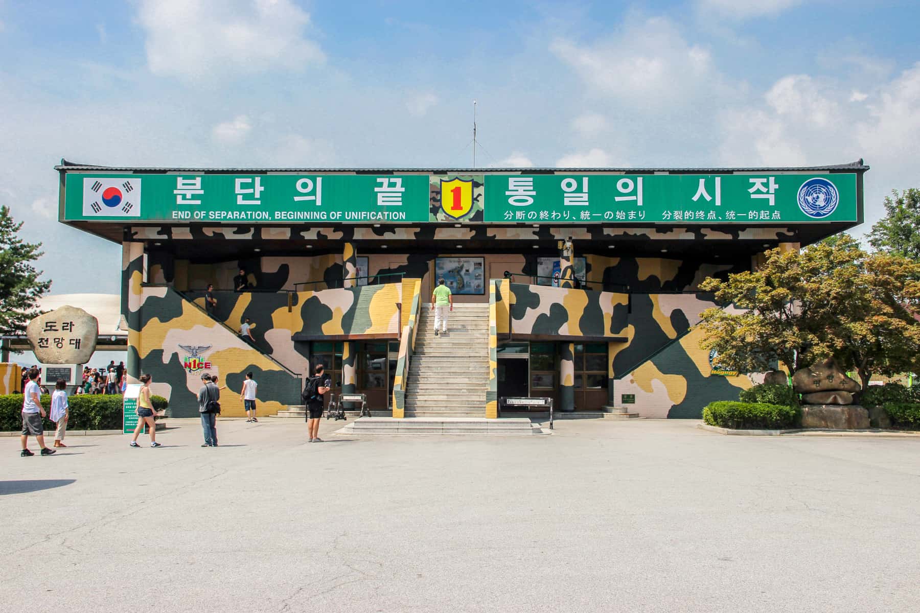 People stand outside a building painted with army camouflage with a green sign with Korean writing - the Dora Observatory at the DMZ.