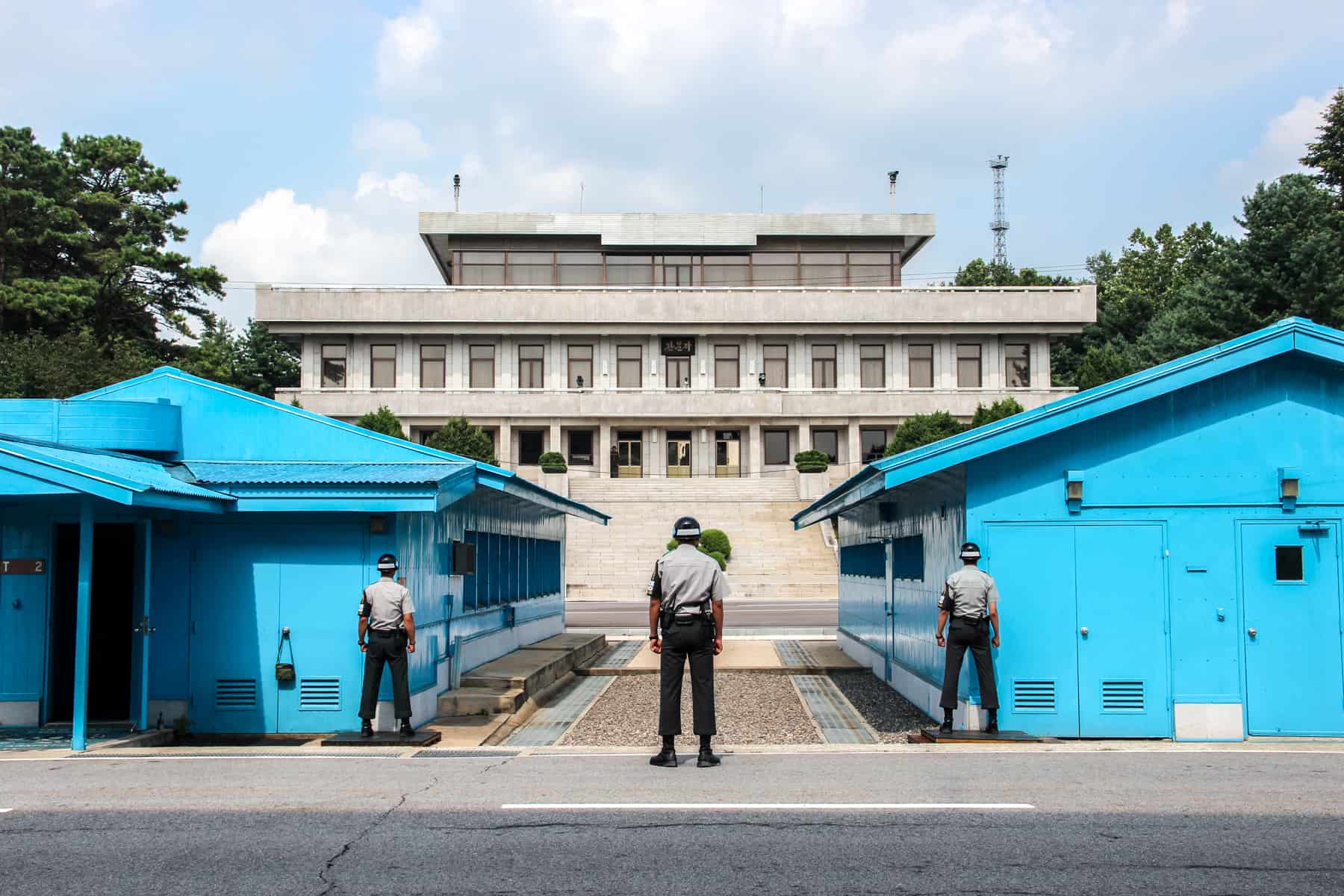 Three South Korean guards in grey and black uniform stand between two blue huts on the North South Korean border at the DMZ demiliterized zone on a DMZ tour. They are facing towards the beige building in North Korea. 