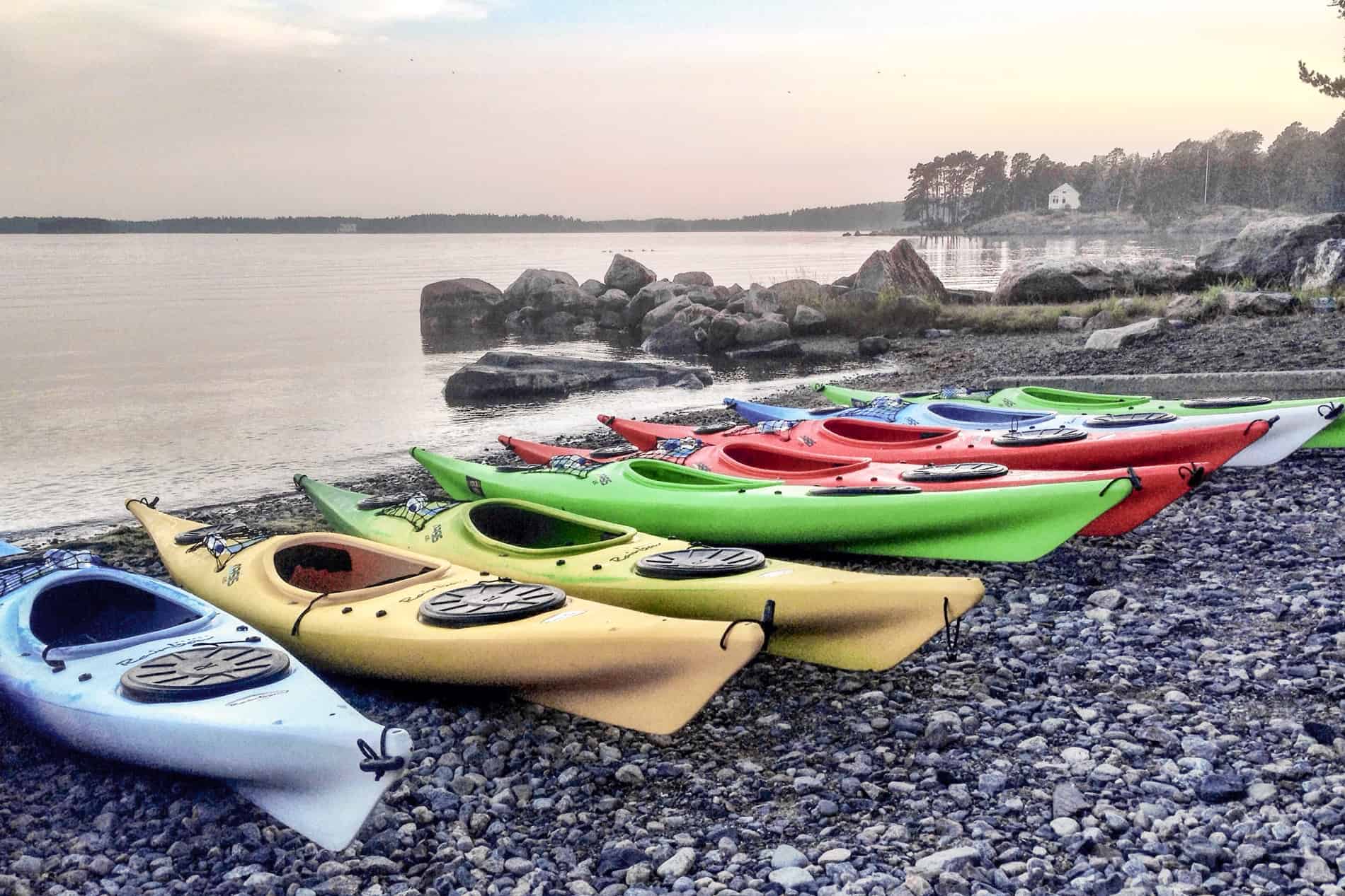 A row of eight blue, yellow, green and red canoes on a pebble beach at a calm water coastline. 