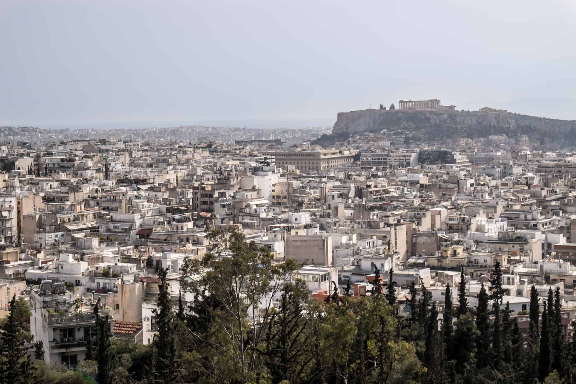 Elevated view over the densely packed buildings of Athens city in front of the Acropolis hill, with the Parthenon structure on top. 