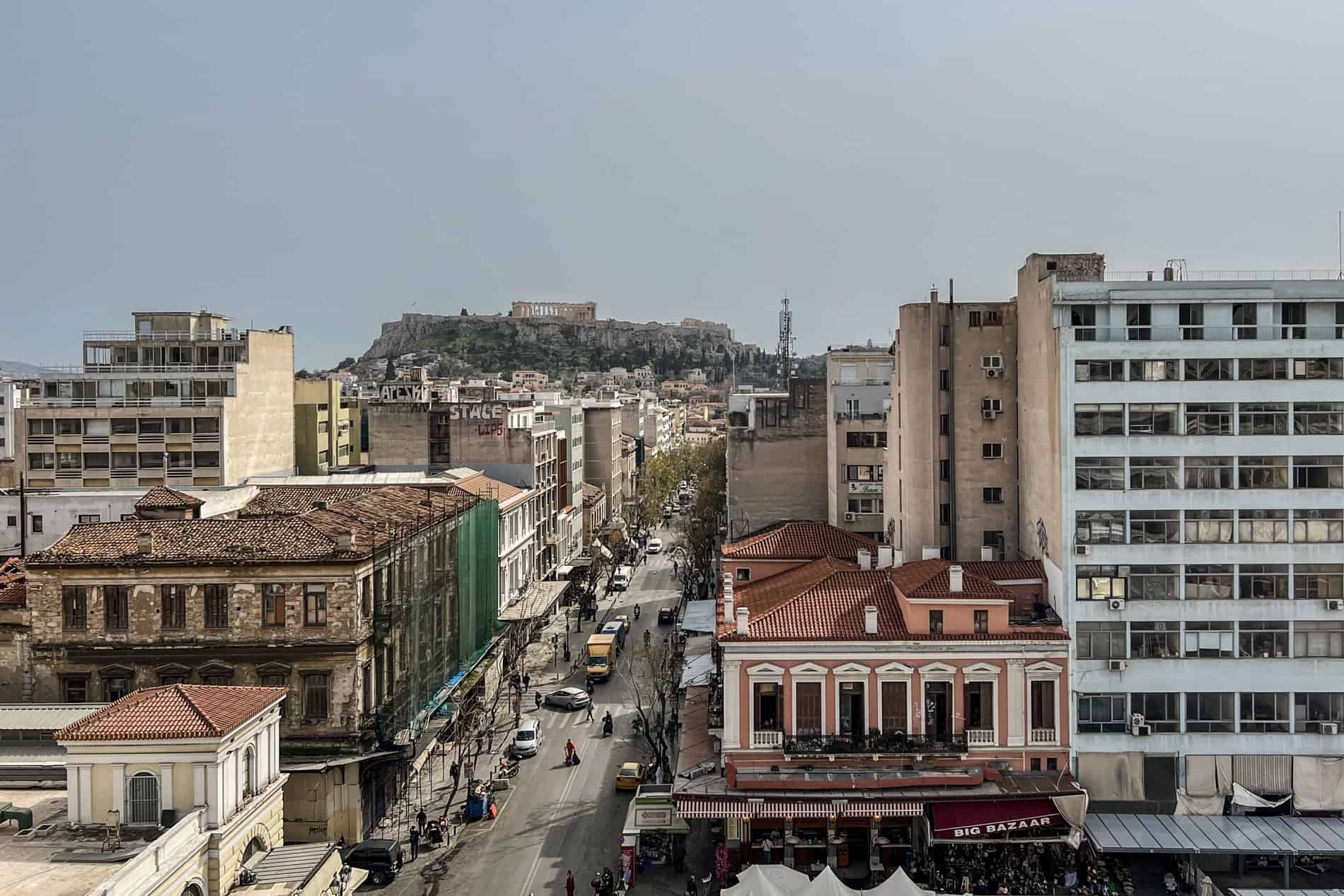 High view to a busy Athens city road lined with modern block buildings, in the direction of the Parthenon topped Acropolis hill.