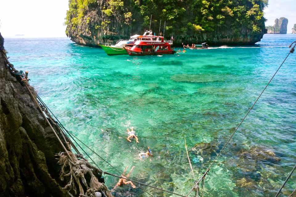 Travellers in blue-green water swim towards a rope ladder to get onto Koh Phi Phi Leh island in Thailand. A red boat waits in the background. 