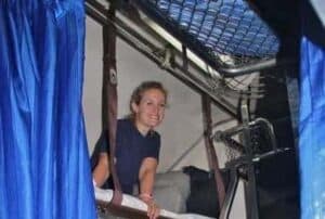 A woman on a train top bunk bed with blue curtains.