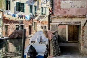 A woman peers around a brick wall on a small street canal in Venice.