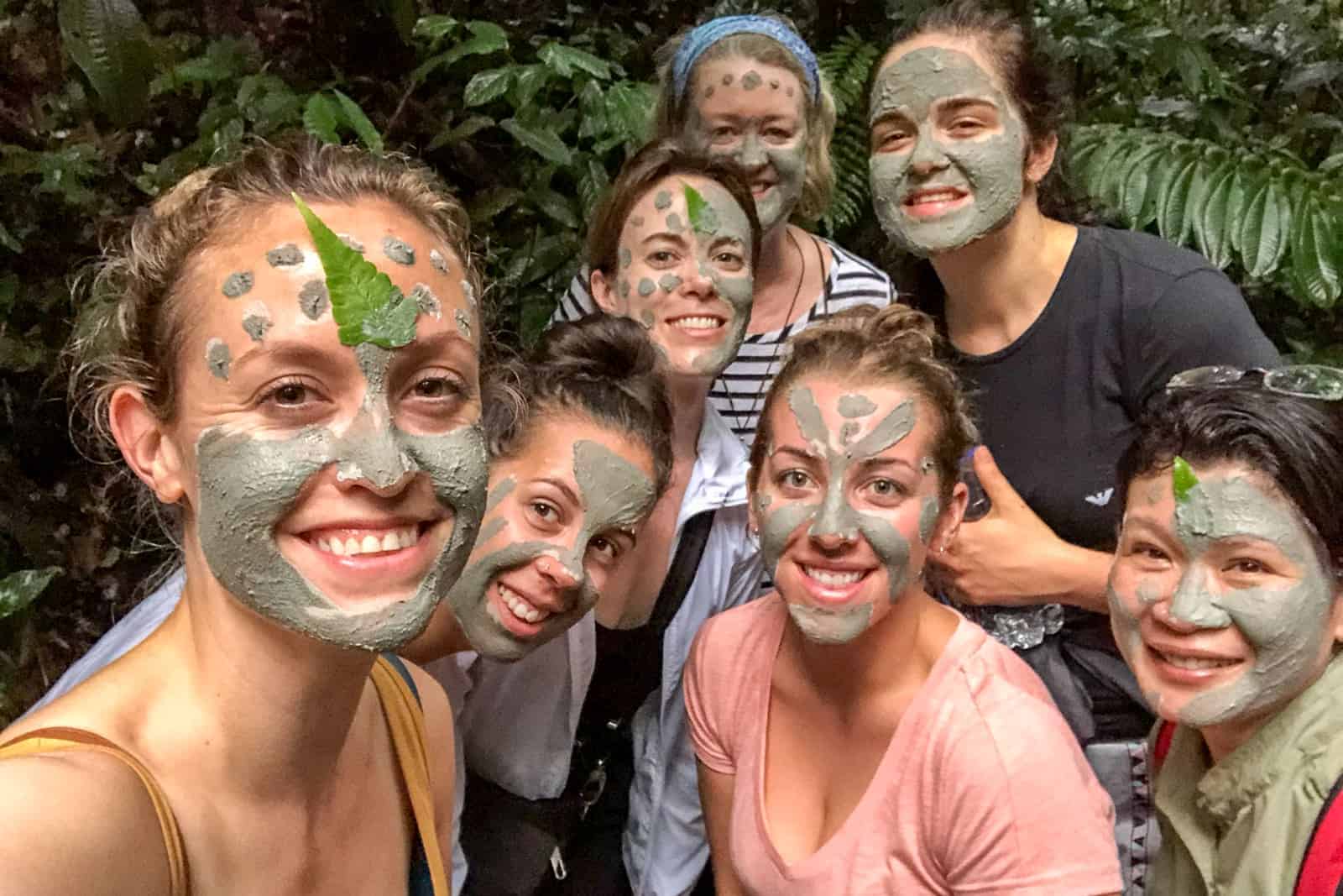 Seven women whose faces are covered in grey, natural clay face masks and leaves, standing in the Ecuador Amazon jungle. 