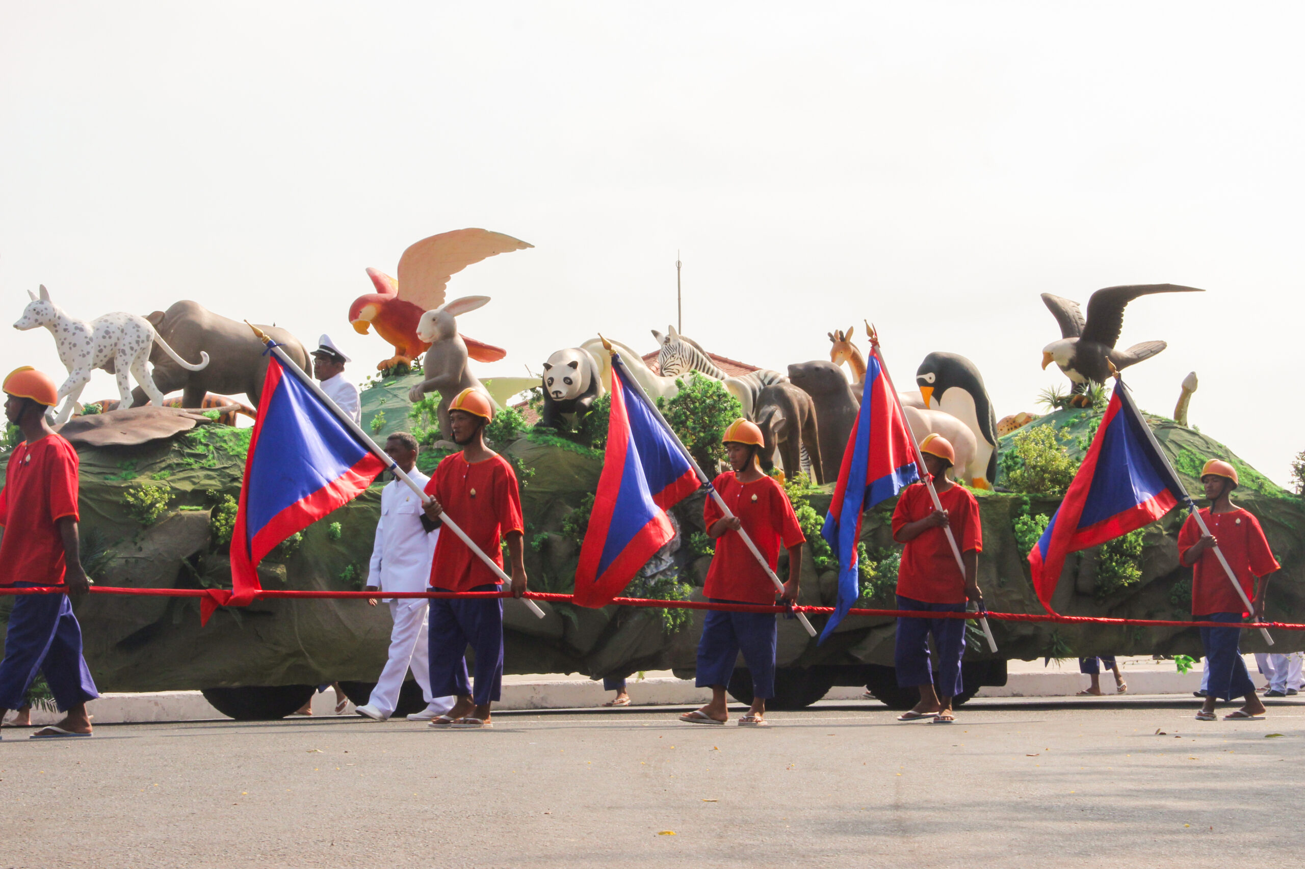 Men in red t-shirts and blue cropped trousers carrying flags in the same Cambodian colours, walk alongside a funeral procession float topped with sculptures of animals including a parrot, a panda, a penguin and an eagle.