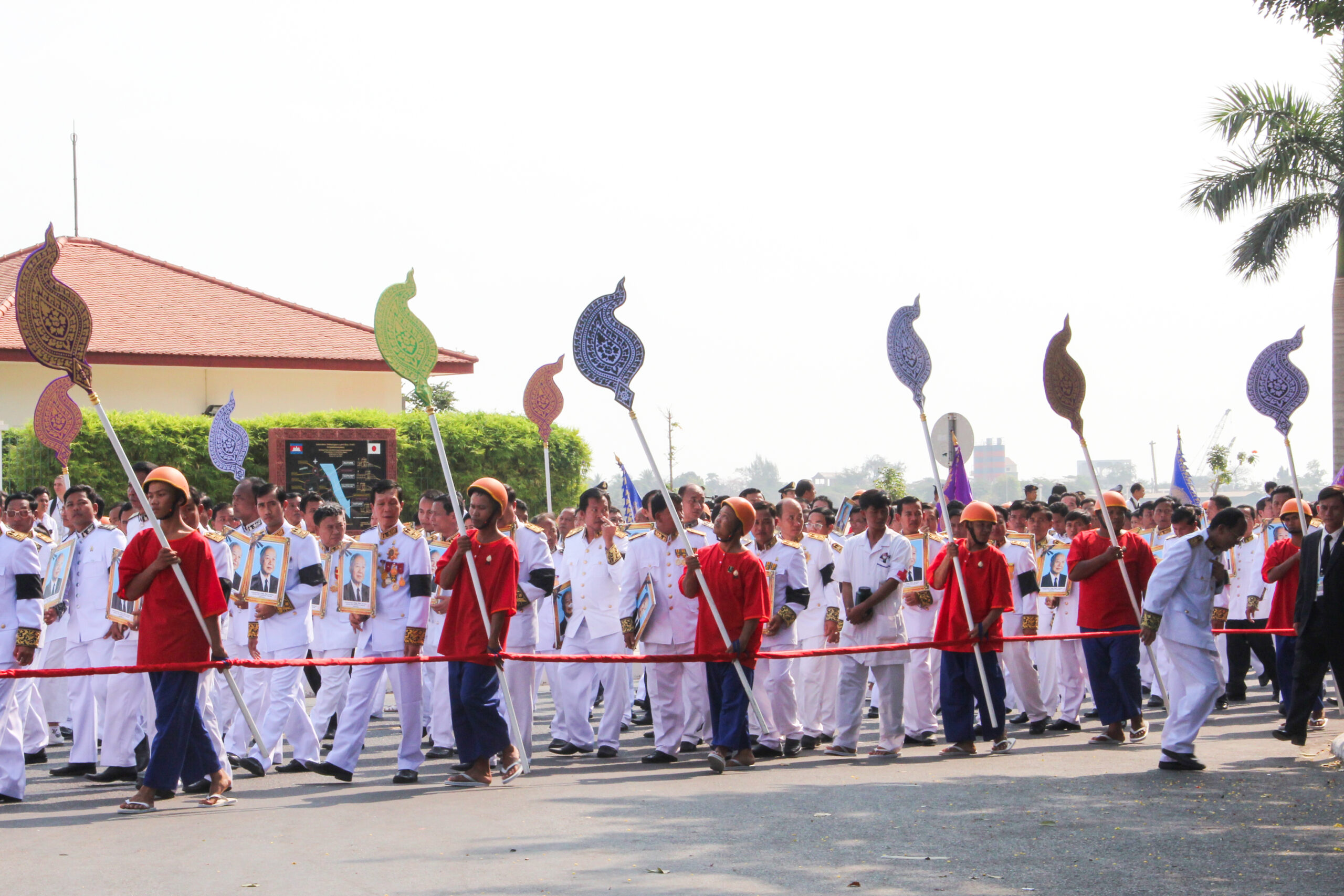 Men in red t-shirts carrying multi-colourded symbols on sticks walk beside a procession of mourners dressed in white carrying a golden framed image of King Norodom Sihanouk in Cambodia.