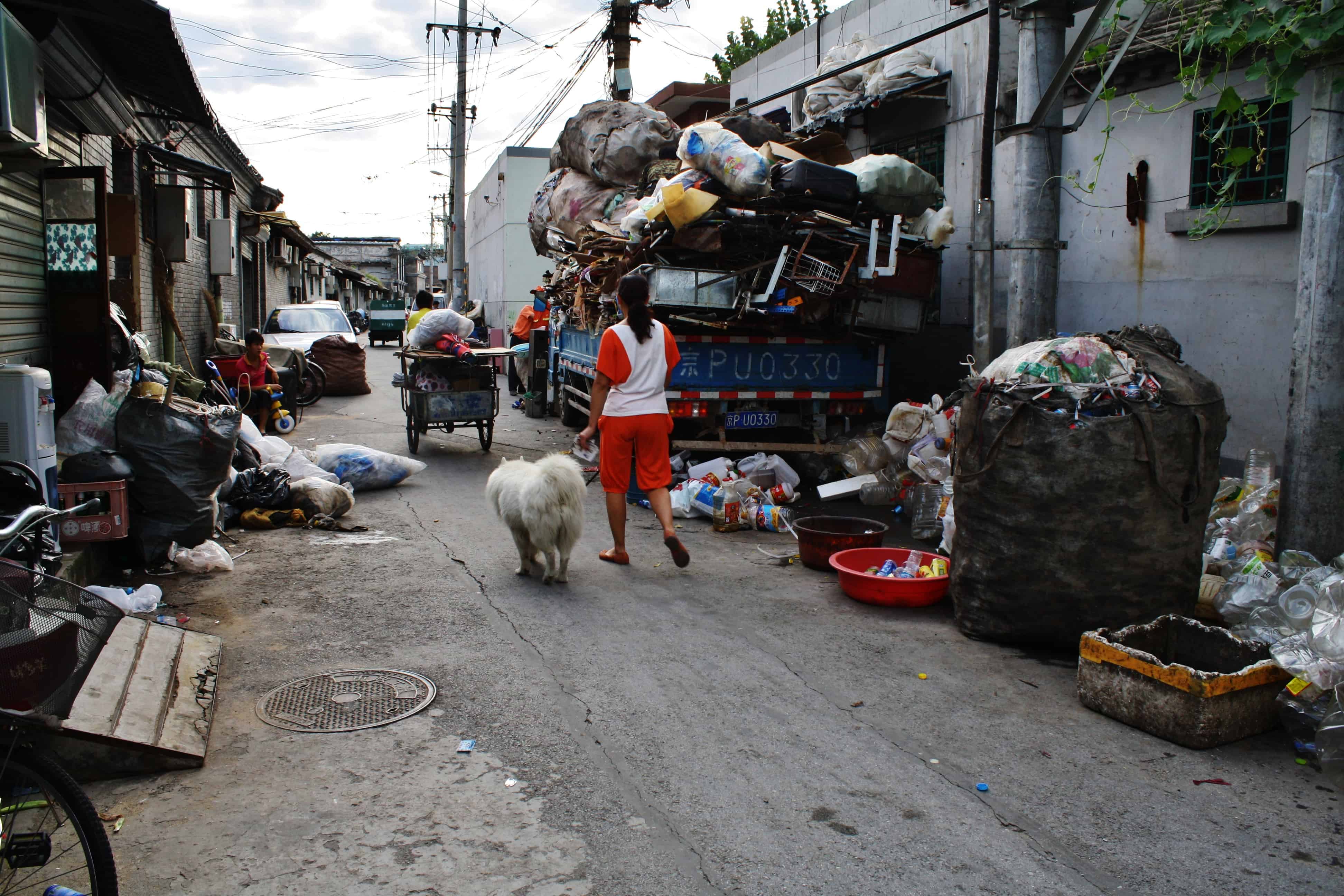 A Woman and her dog walking on a street filled with litter in a Hutong in Beijing