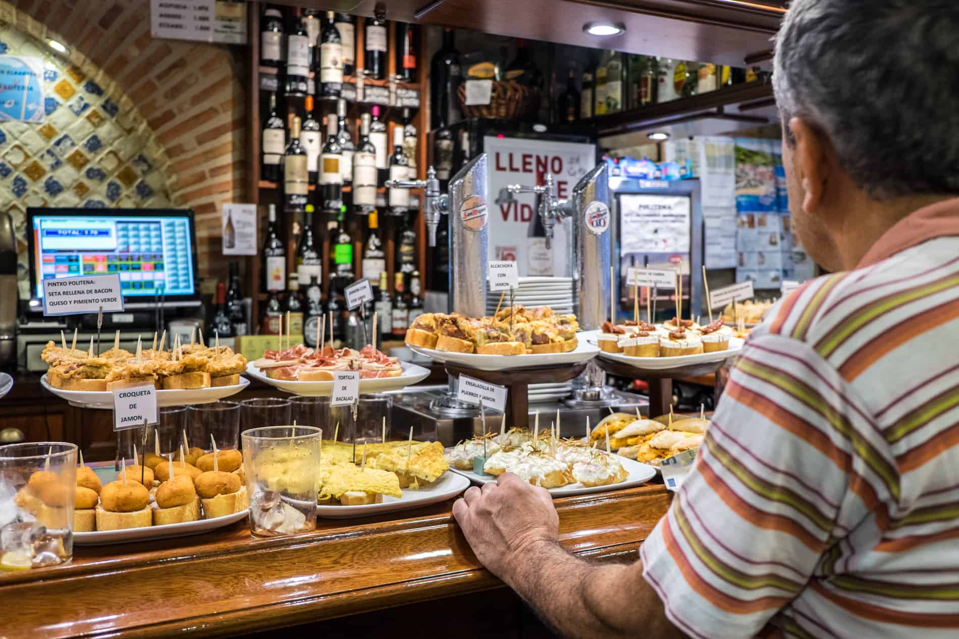 A man faces a dark wooden bar covered in plates of the best Pintxos in San Sebastian – snacks on cocktail sticks. 