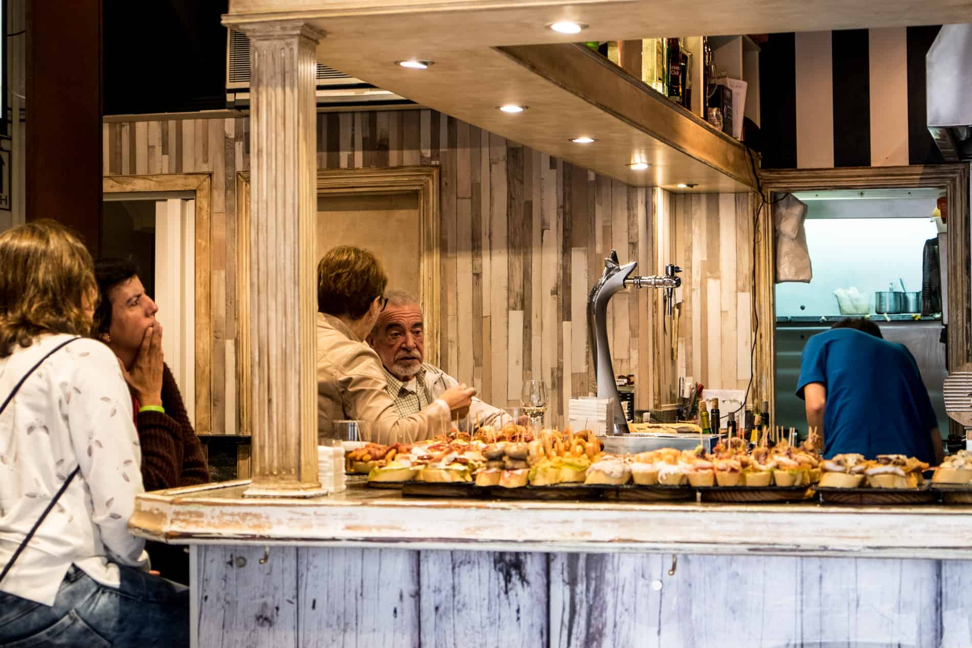 Four people at a bar in San Sebastian that covered in small snacks known as Pintxos. 