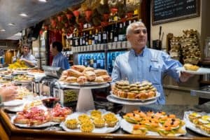 A man in a blue shirt hands over a plate of food across a bar filled with multiples plates of Pintxos bar snacks.