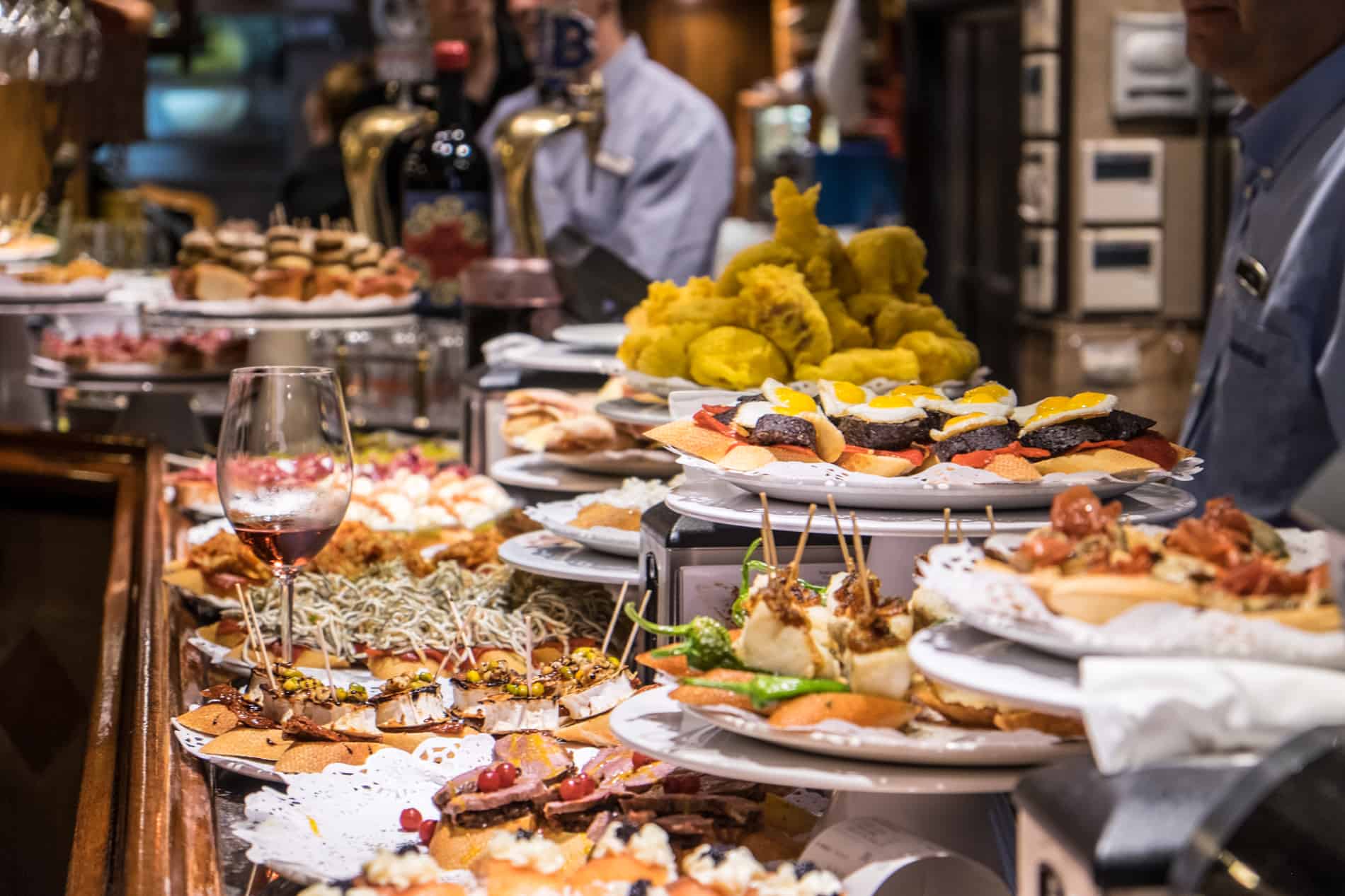 A glass of red wine stands next to plates of bitesized bar snacks, which completely cover a bar in San Sebastian, Spain.