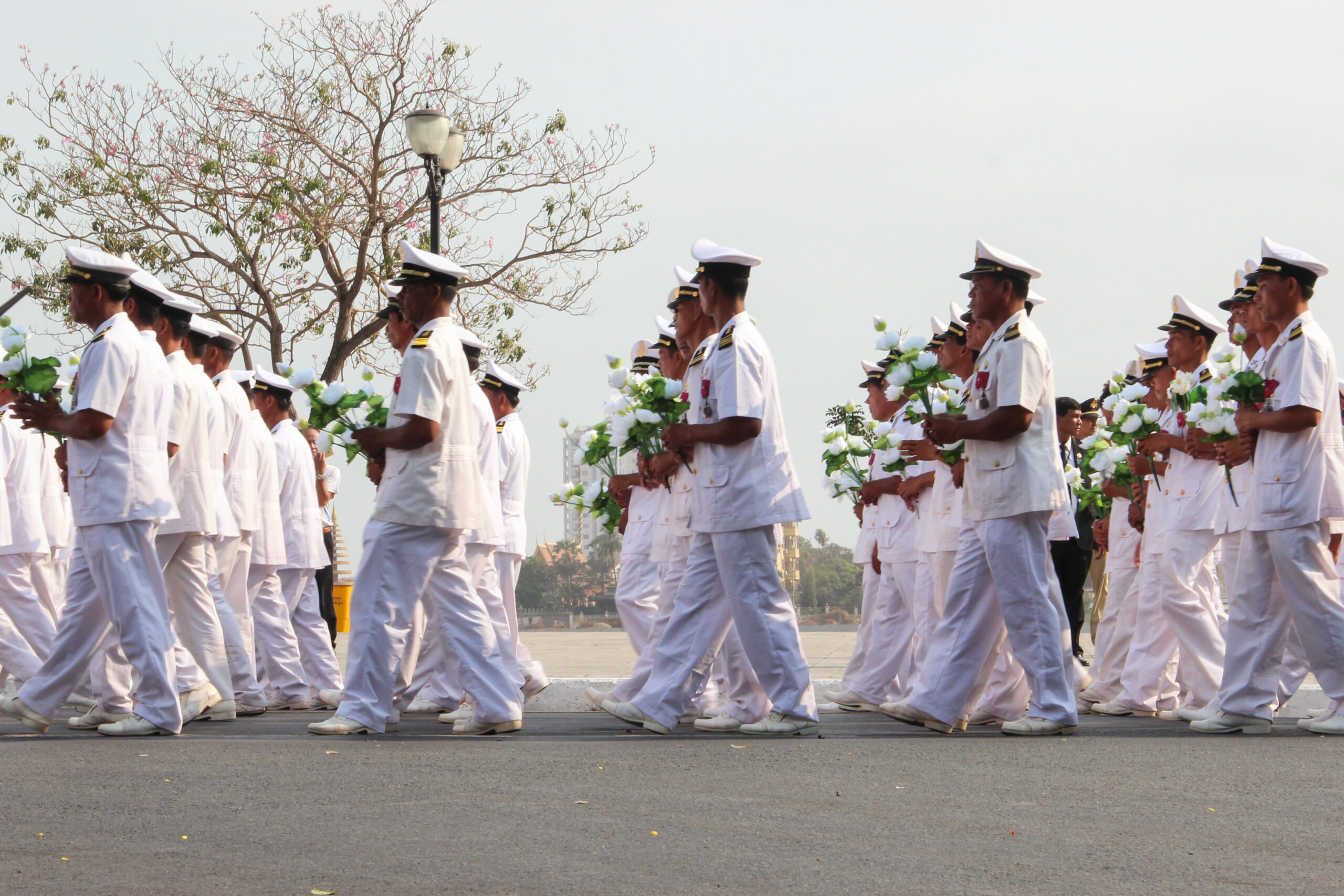Cambodian navy officers dressed in white and carrying white flowers walk in unison alongside a river in Phnom Penh during a state funeral.