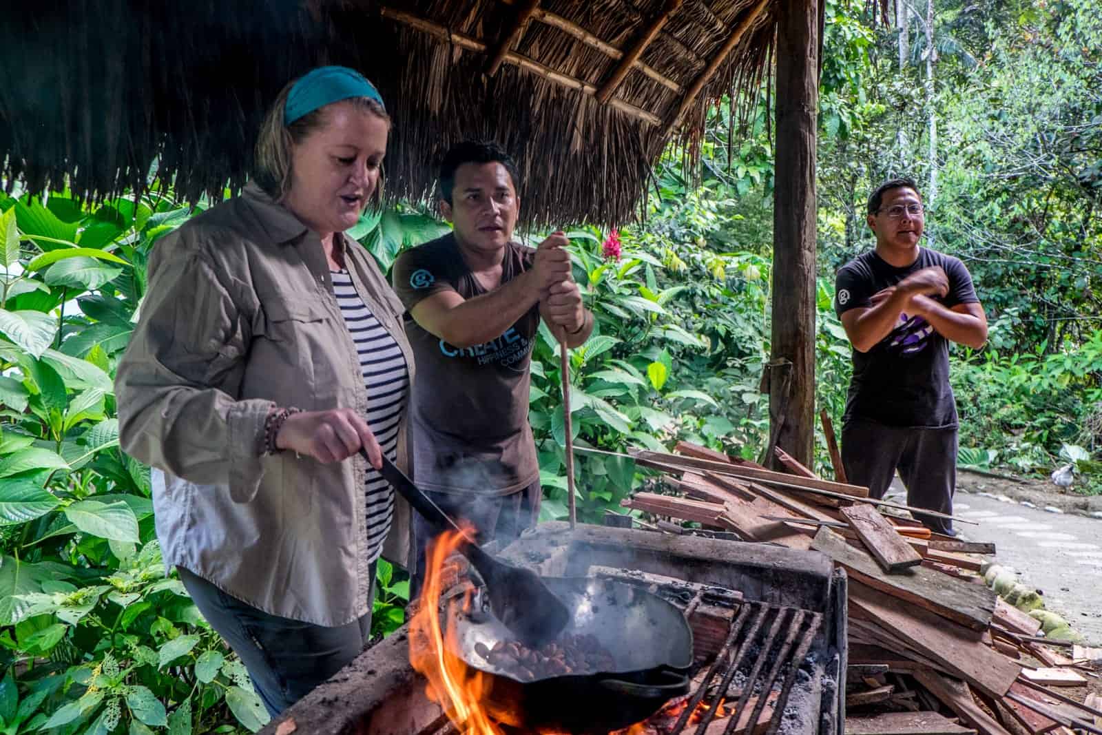 A woman stirs an iron pan full of cacao beans on an open fire grill. She stands under a thatched roof with two men, surrounded by the green of the Ecuador Amazon jungle.