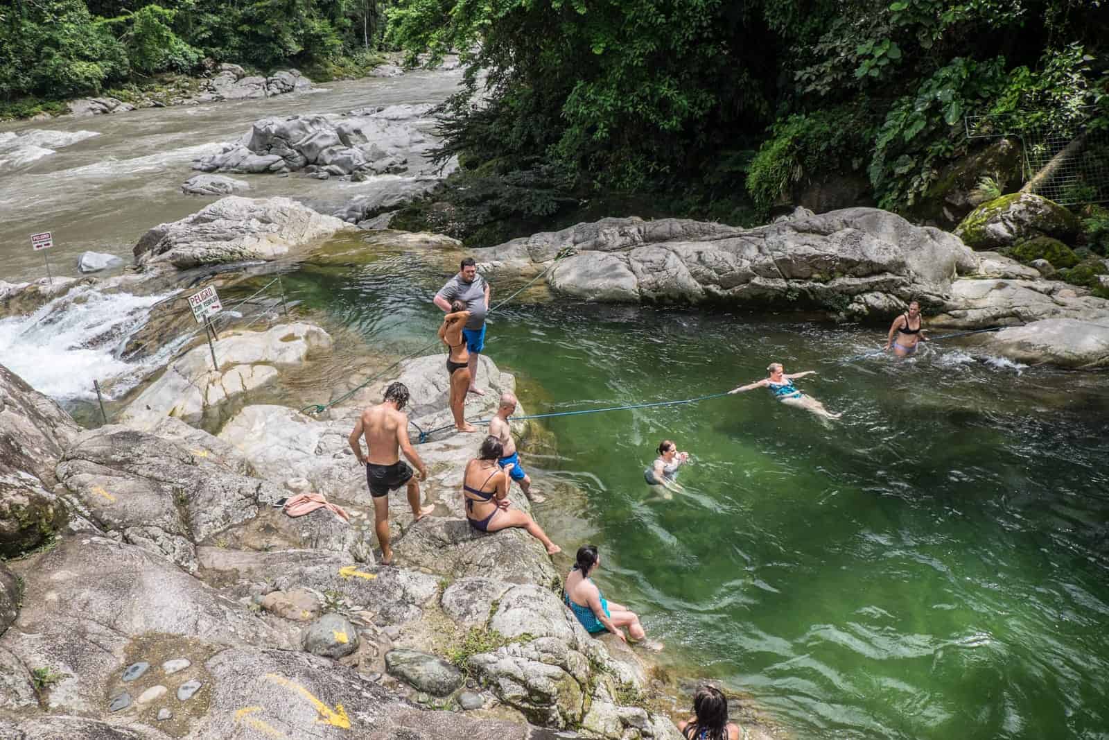 A group of people stand and sit on large rocks while others swim in the green waters of a lagoon in the Ecuador Amazon Rainforest.