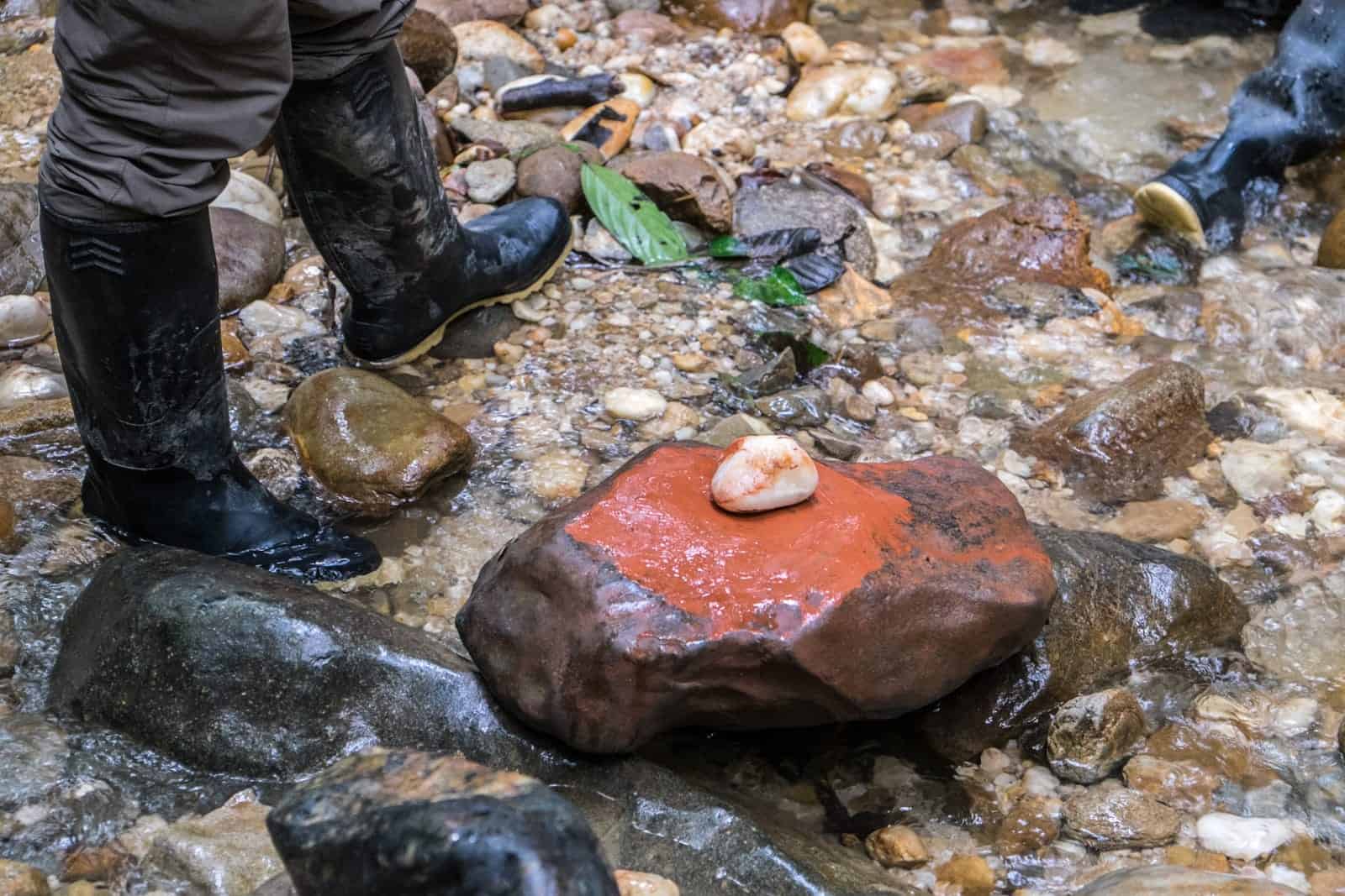 A person in black boots stands next to a large stone, covered in natural red clay dye with a small white stone on top.