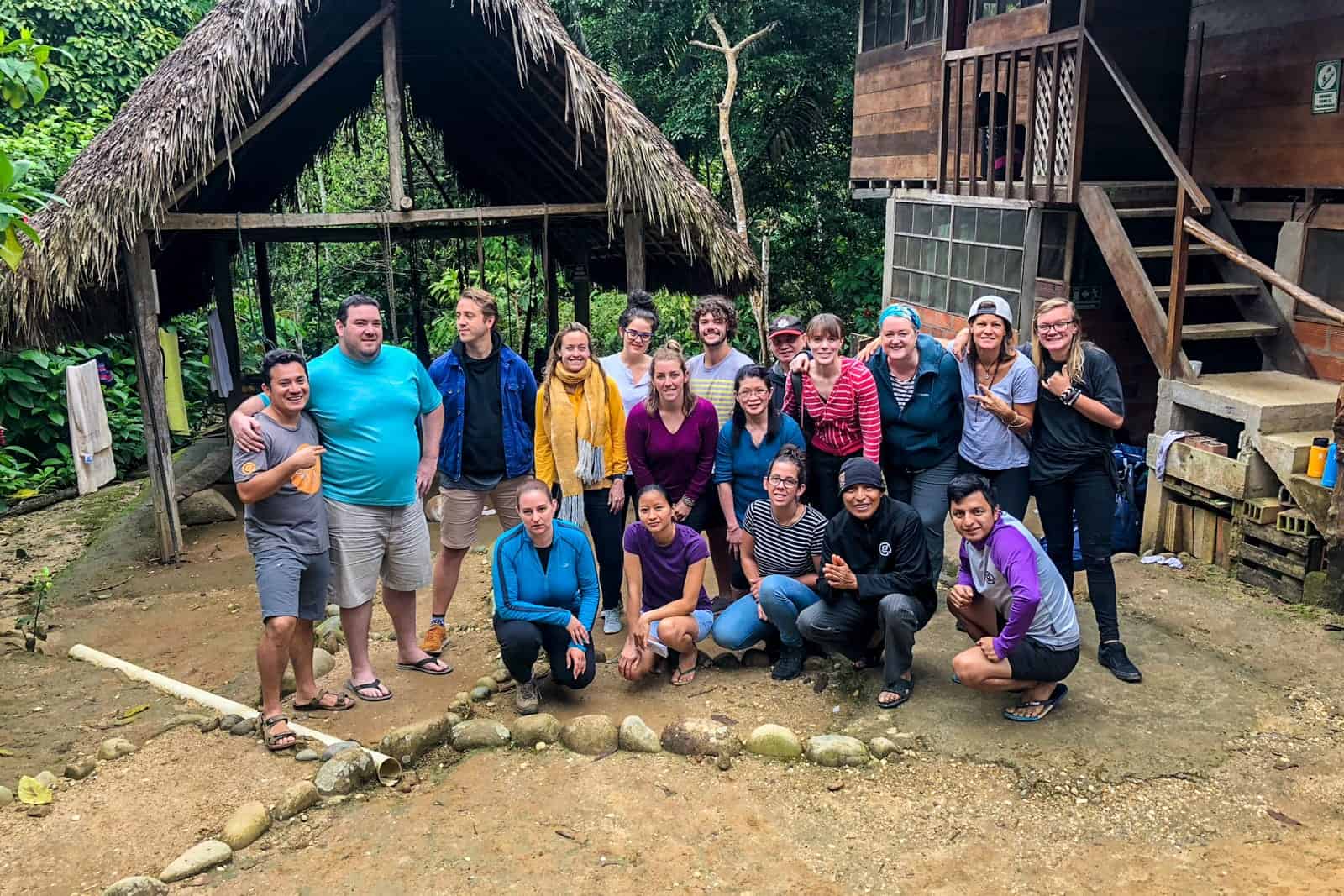 A group of people pose for a group shot next to a wooden house and a backdrop of jungle - their local living lodge in the Ecuador Amazon.