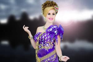 A woman with a blonde and brown hair piece and heavy makeup, poses in a traditional Khmer outfit made from purple fabric embossed with gold chains and gold and silver beading, at a Cambodian photo shoot.