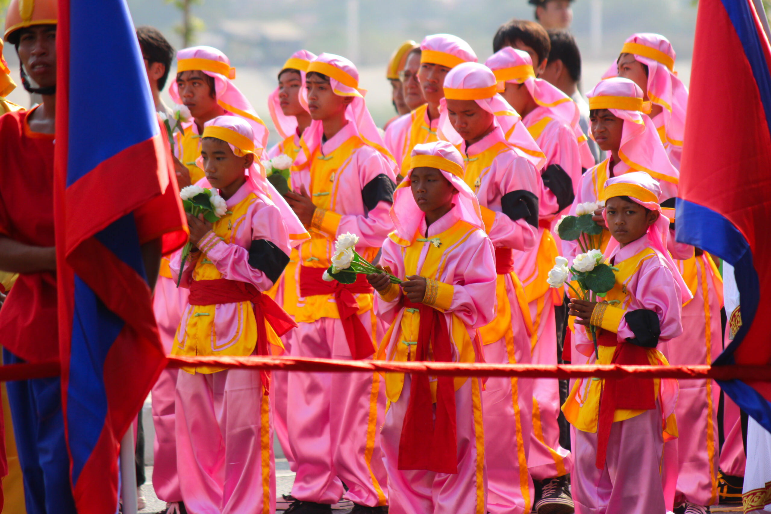 A group of children dressed in bright pink and yellow outfits and carrying white flowers parade alongside Cambodian flags during the state funeral of Norodom Sihanouk.