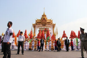 A parade of people walk alongside a tall, golden temple structure with a boat shaped base, carrying former Cambodian King Norodom Sihanouk in Phnom Penh at his state funeral.