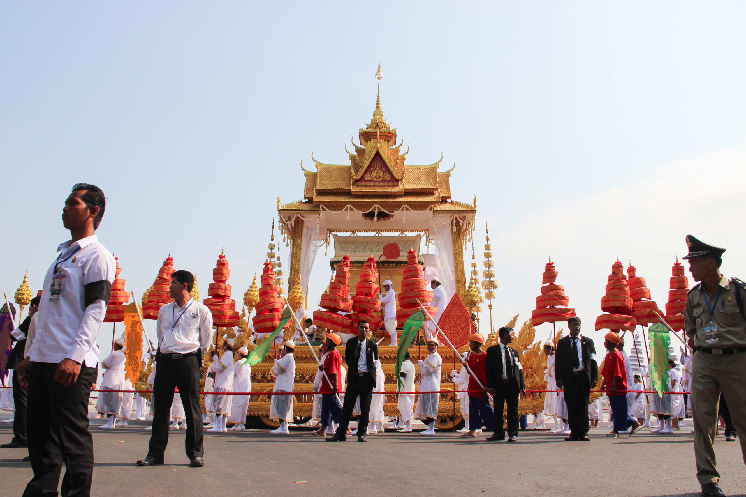 A group of people dressed in white and carrying orange decorations walking alongside a grand golden float carrying the golden coffin sarcophagus of Norodom Sihanouk, King of Cambodia during his state funeral.