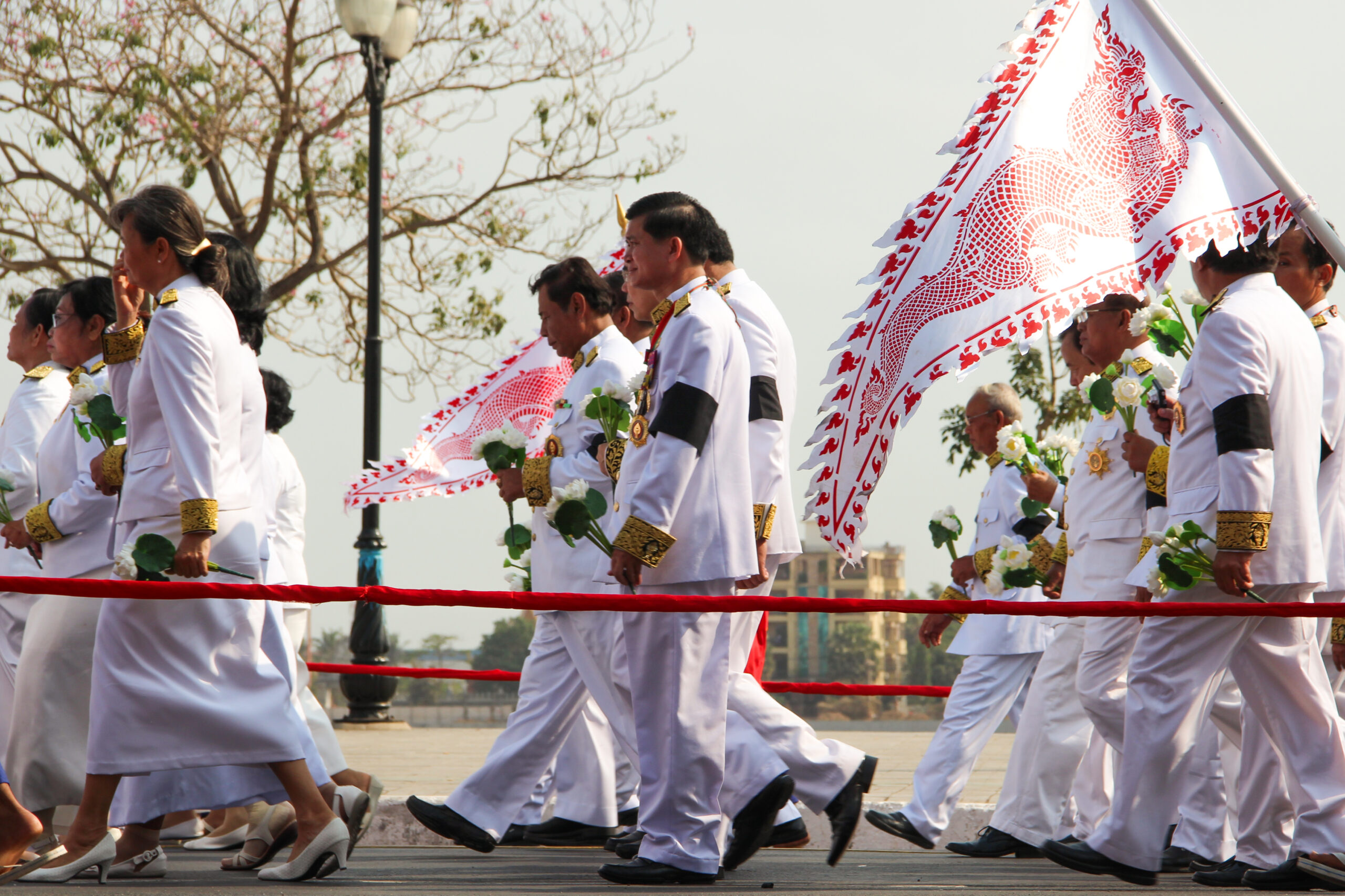 A group of people dressed in white (the men with black armbands) parade alongside a river with white and red flags during a state funeral in Cambodia.