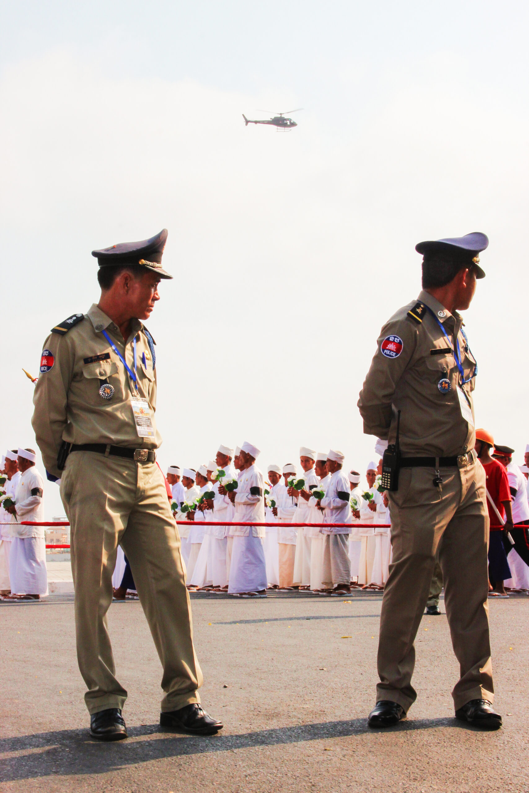 Two Cambodian policeman keep guard as a group of people dressed head to tow in white parade behind. A helicopter can be seen in the air.