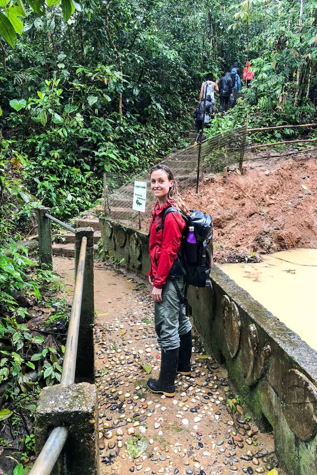 A woman in a red jacket, drenched from the rain, walks through a stone pathway towards some steps that lead further into the Amazon Rainforest in Ecuador. 
