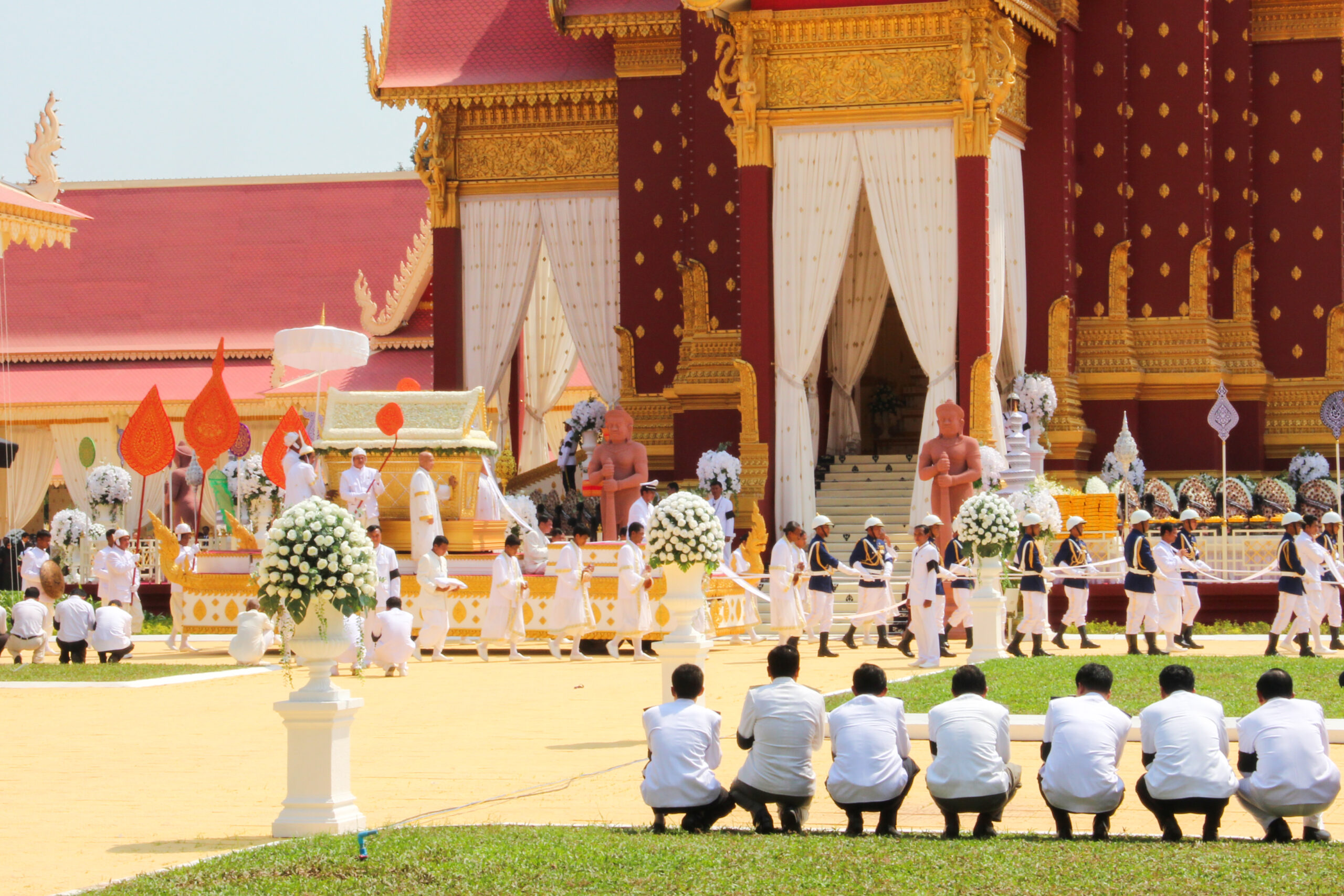 In the Phnom Penh royal palace grounds, men in white shirts and black trousers crouch down in front of a parade of people also dressed in white, carrying a golden coffin.