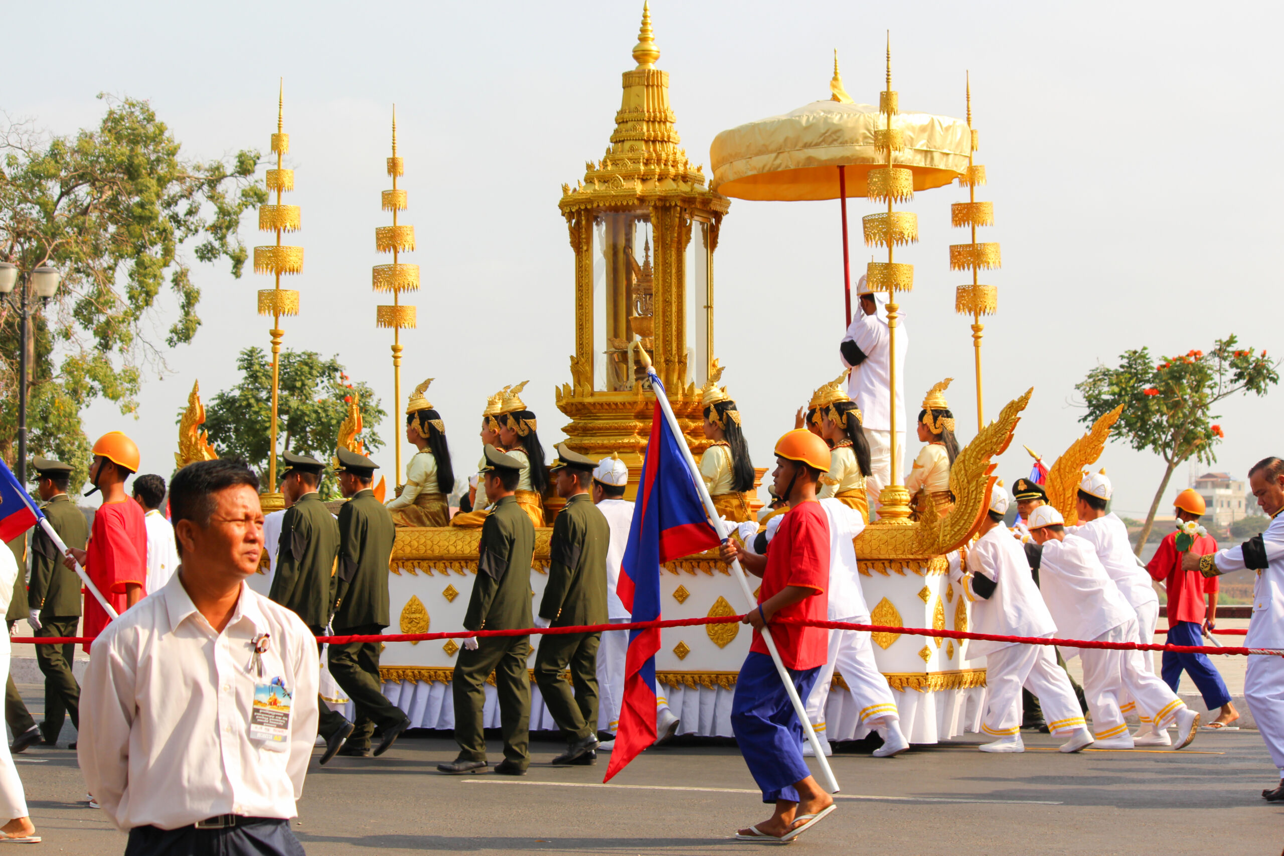 A parade of people dressed in white and khaki green pushing a float adorned with a golden temple-like structure and a dozen seated women with golden headwear.