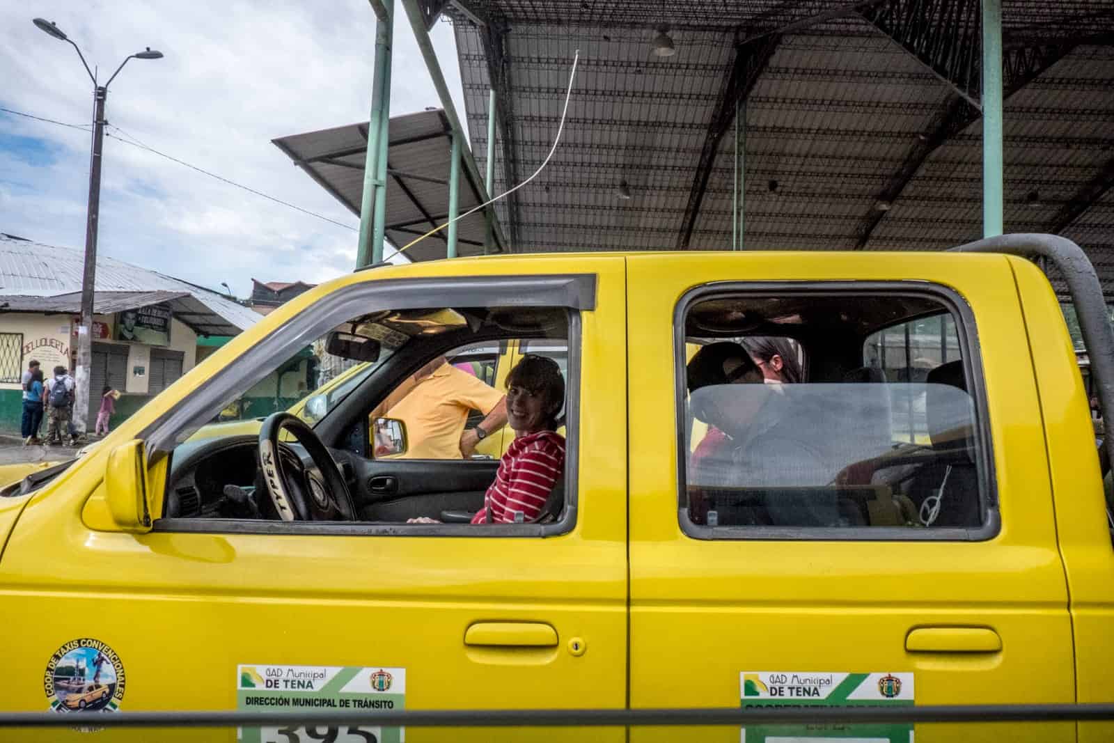 A woman smile in the passenger seat of a yellow Taxi in Tena, Ecuador. Other passenger can be seen squeezed into the back seat. 